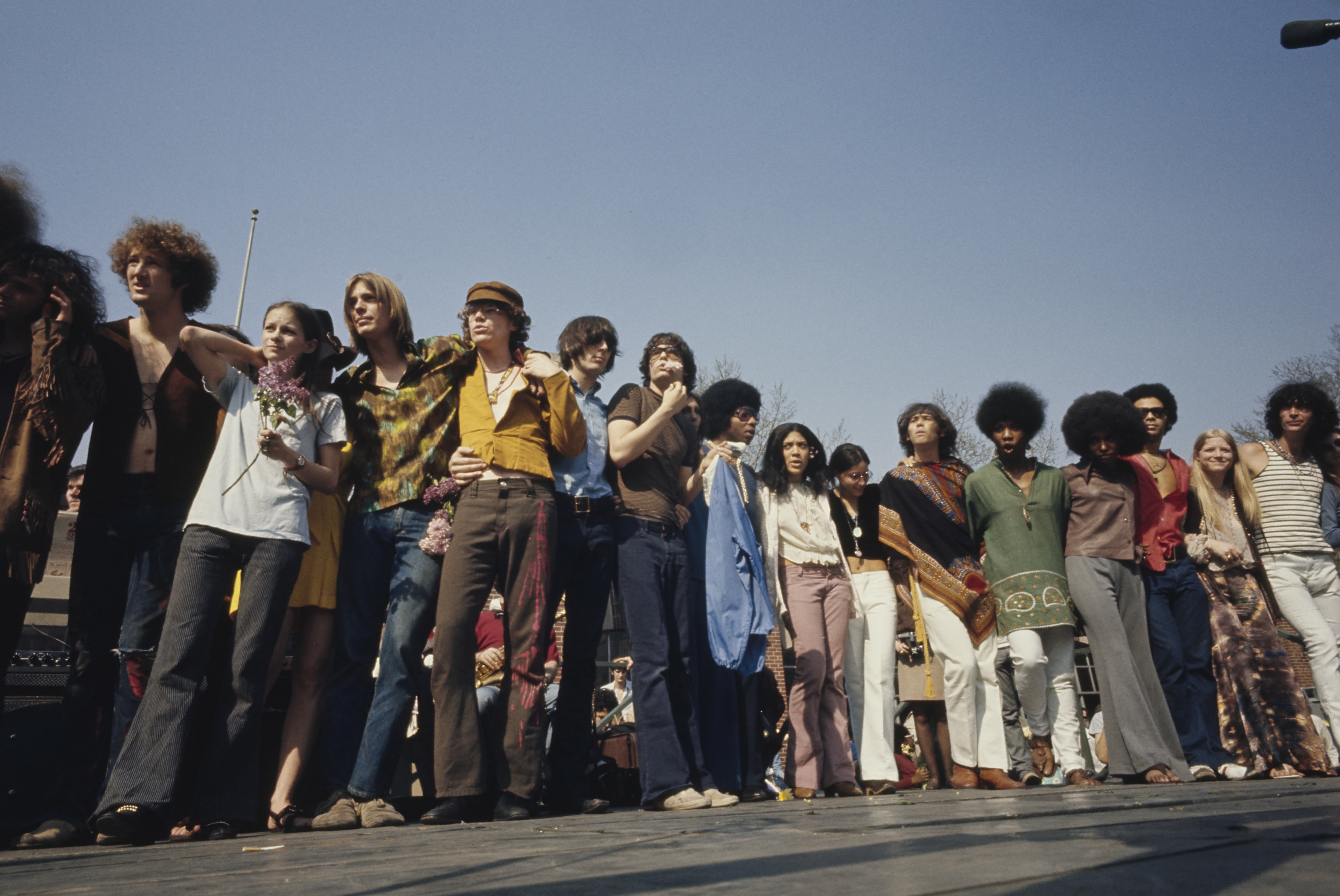 Group of people, including men and women, standing arm in arm at an outdoor event, dressed in 1970s, bohemian-style clothing