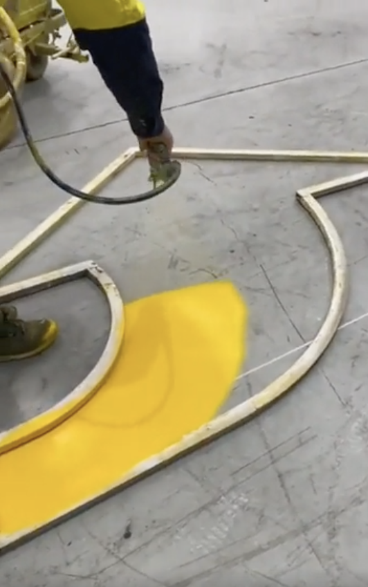 A person is seen spray painting with yellow paint using a stencil on what appears to be a pavement or ground surface