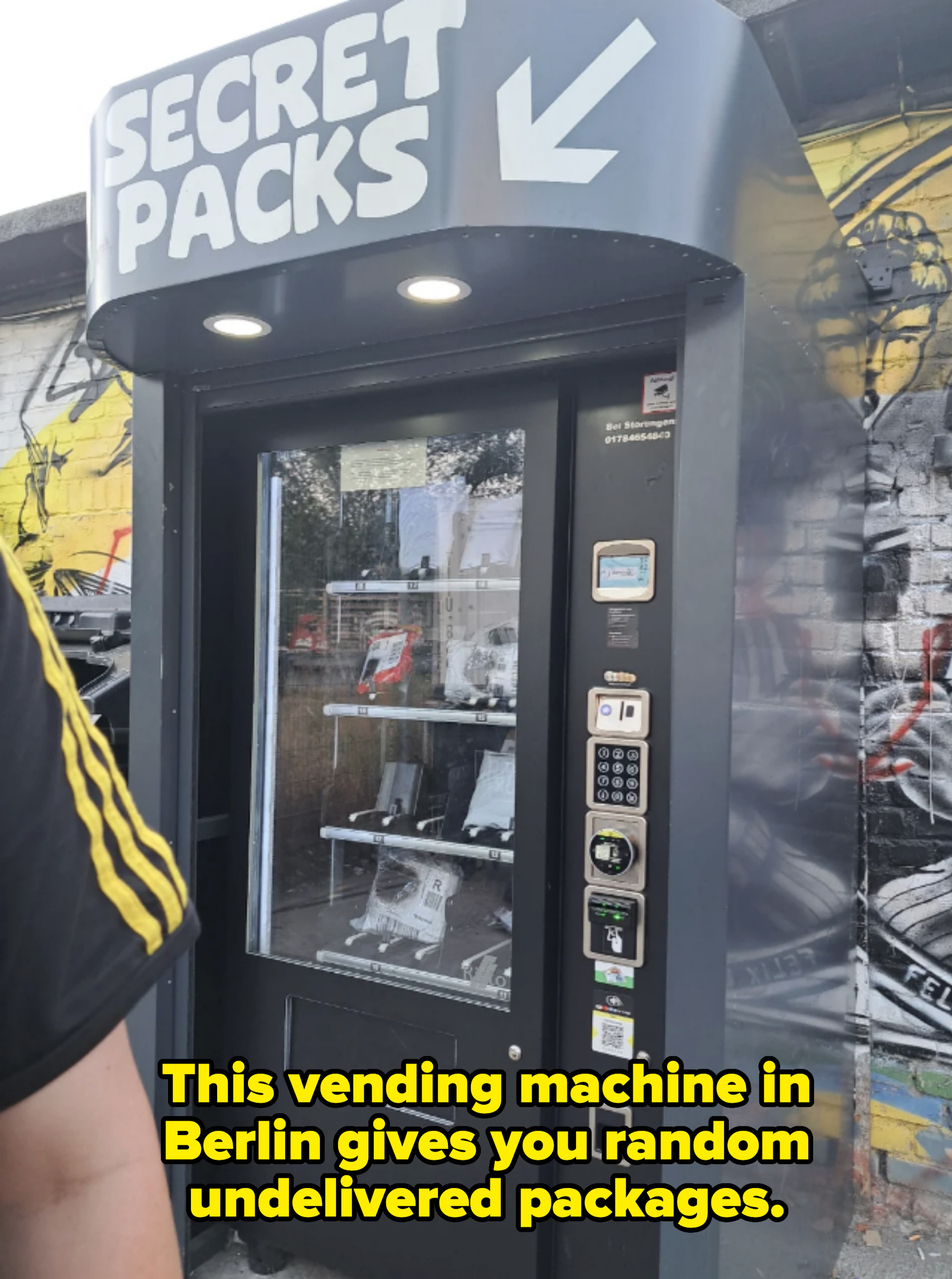 Photograph of a vending machine labeled "Secret Packs" with various control buttons and a digital screen, set against an urban graffiti wall