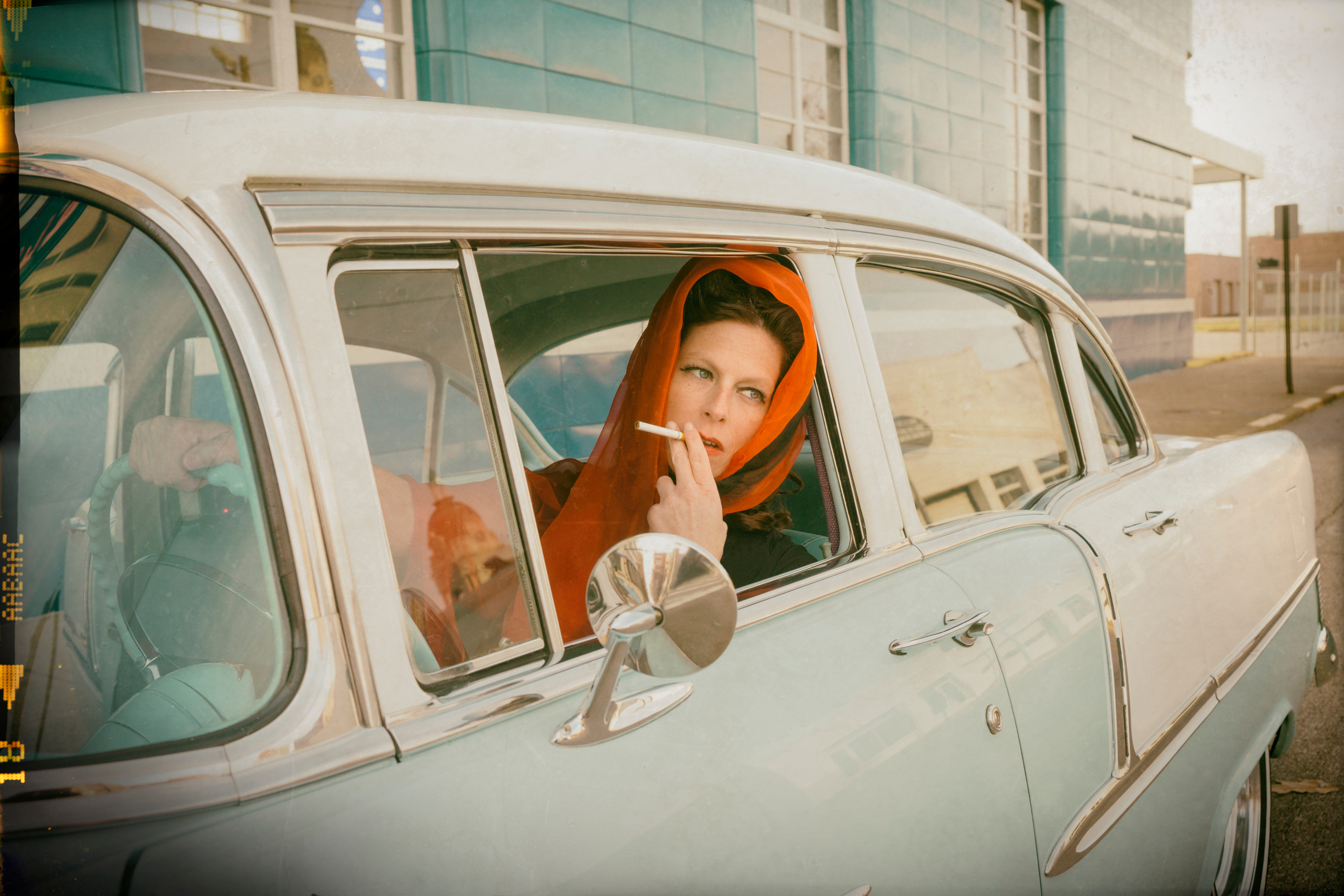 A woman wearing a red headscarf sits in a vintage car, holding a cigarette and looking out of the open window