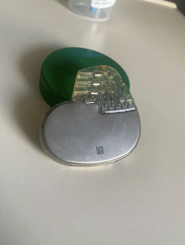 A medical device, resembling a pacemaker, rests on a table next to a green object that could be a container lid