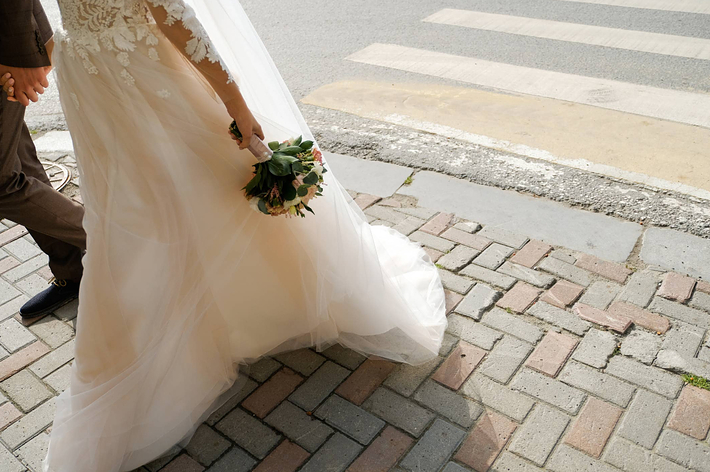 新郎新婦が手を繋ぎ、花嫁がウェディングドレスを着て歩道を歩いている。花嫁はブーケを持っている。