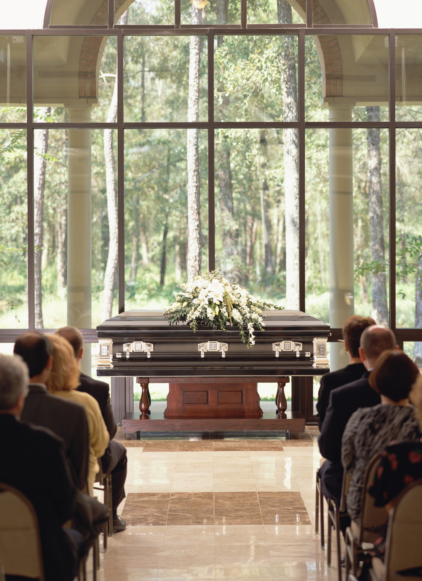 A funeral service with attendees seated, facing a casket adorned with flower arrangements, located in a room with large windows showing trees in the background