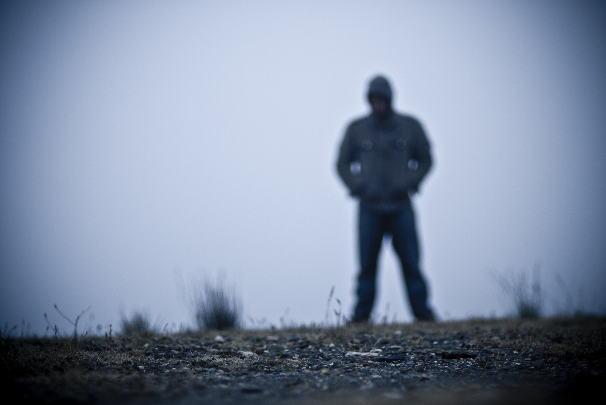 A blurred figure stands on a desolate, foggy landscape with hands in pockets, facing the camera. The figure's features and details are unrecognizable