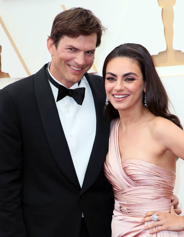 Ashton Kutcher and Mila Kunis smile together on a red carpet. Ashton wears a tuxedo, and Mila wears a one-shoulder gown