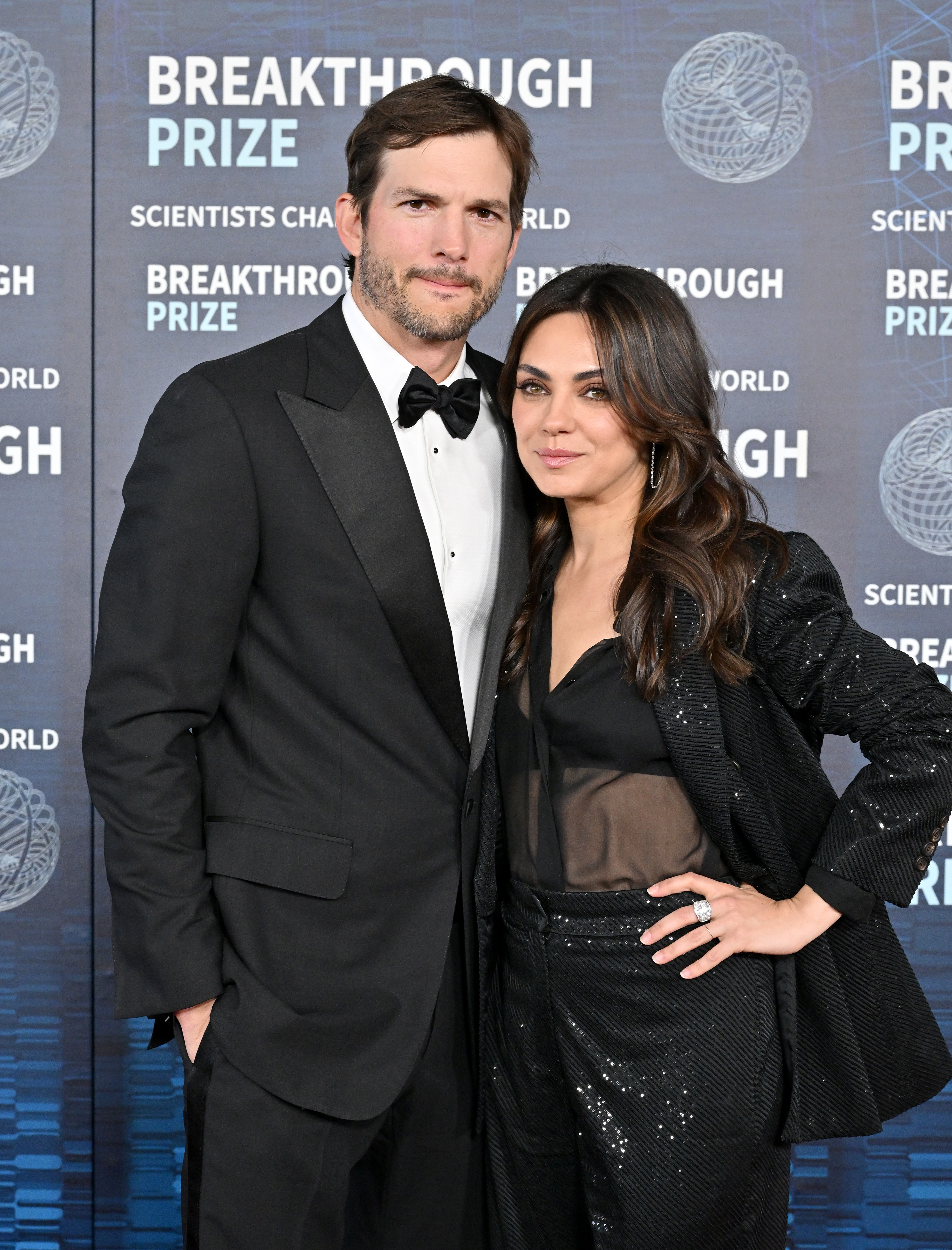 Ashton Kutcher and Mila Kunis at the Breakthrough Prize event, posing together. Ashton is in a tuxedo, and Mila is wearing a stylish pantsuit