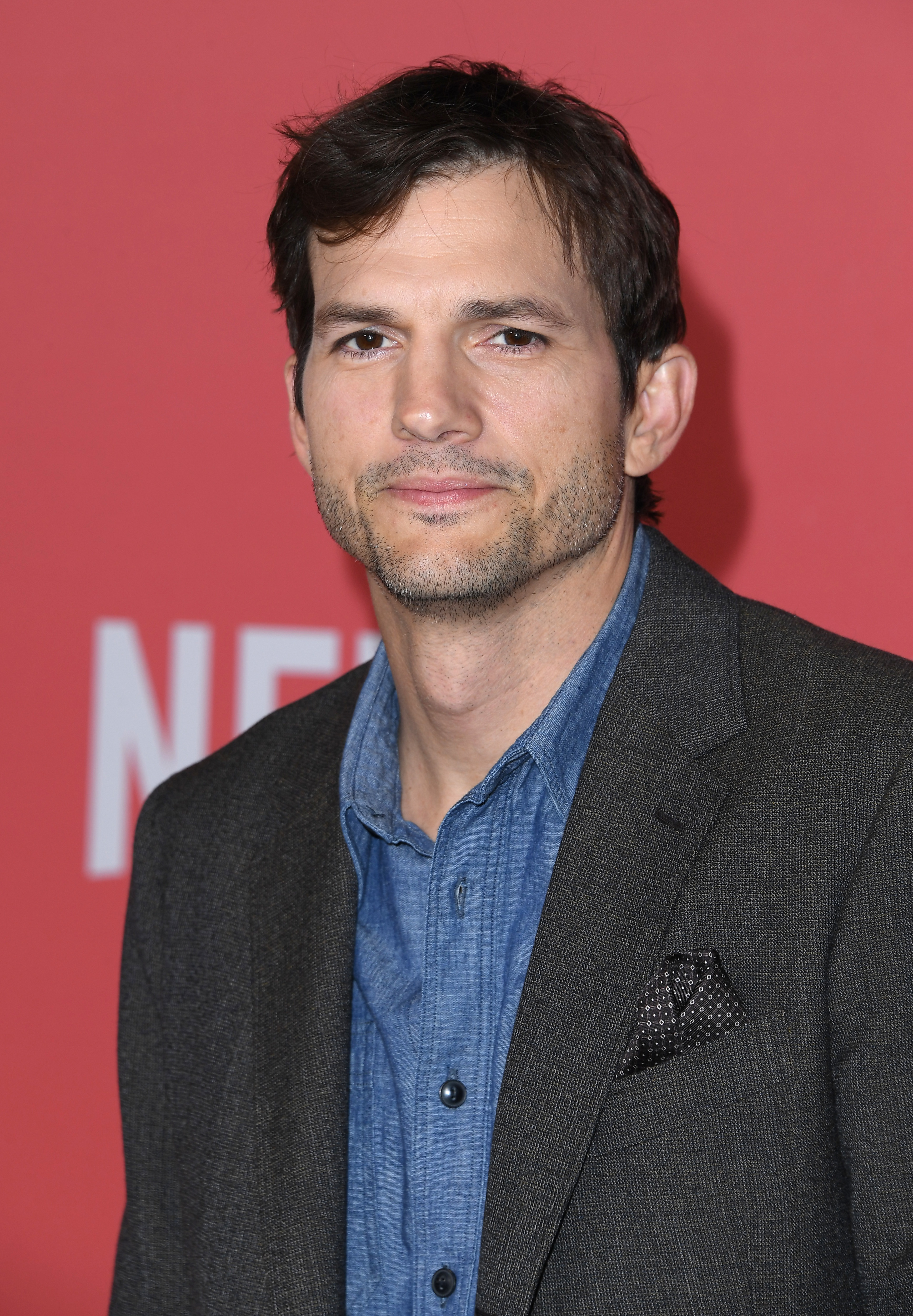 Ashton Kutcher in a gray blazer over a blue shirt poses in front of a backdrop with the Netflix logo