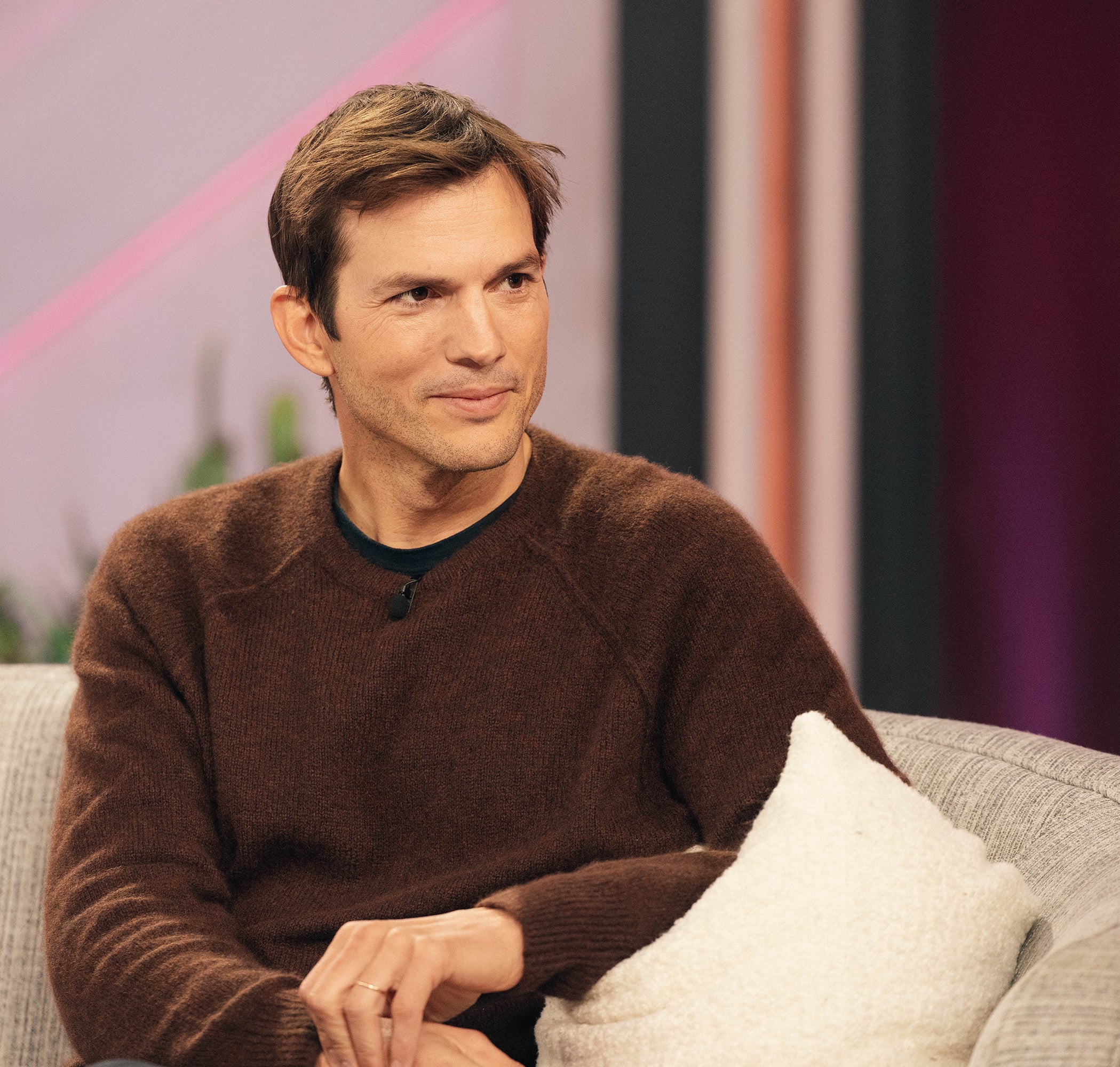 Ashton Kutcher smiles while sitting on a couch, wearing a casual sweater, on a talk show set