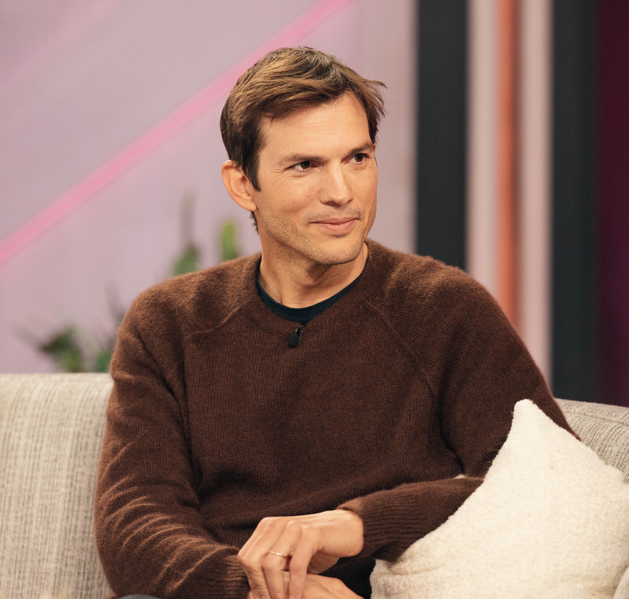 Ashton Kutcher smiles while sitting on a couch, wearing a casual sweater, on a talk show set