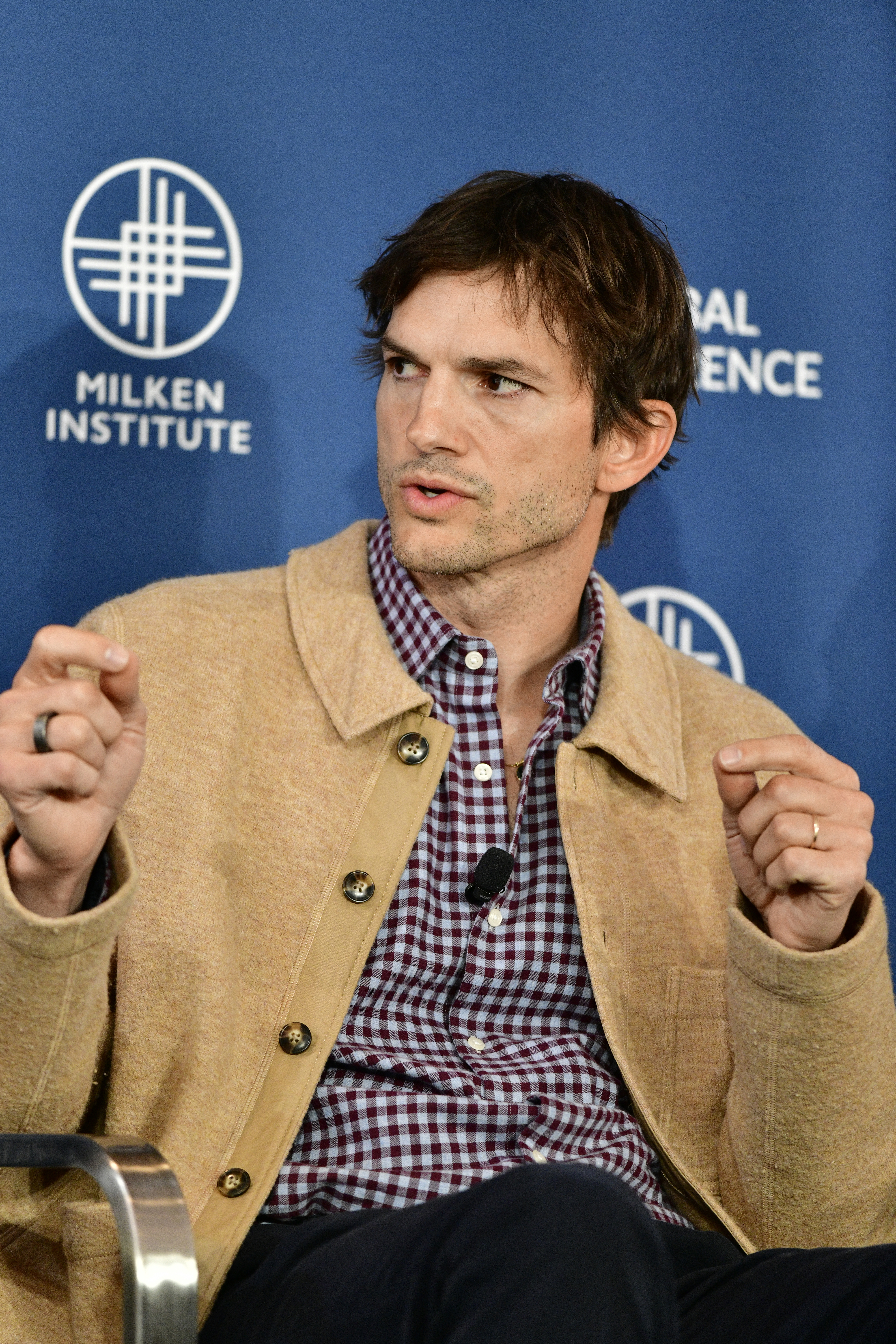 Ashton Kutcher speaks at a Milken Institute event, wearing a checkered shirt under a jacket