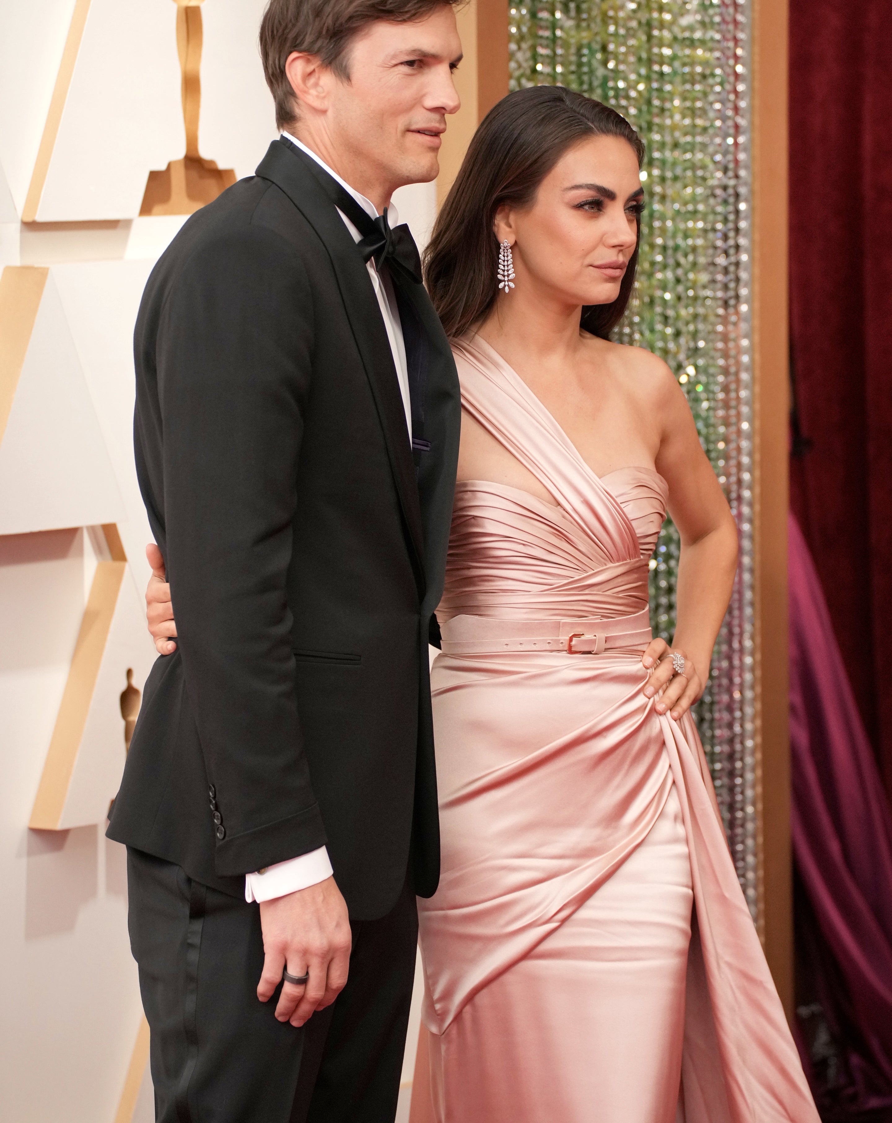 Ashton Kutcher in a tuxedo and Mila Kunis in an elegant one-shoulder gown pose together on a red carpet