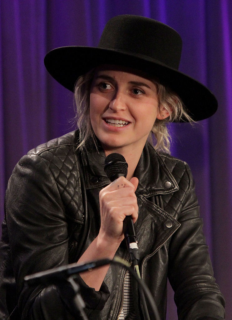 Sarah Jones, wearing a black leather jacket and wide-brimmed hat, holds a microphone during an interview