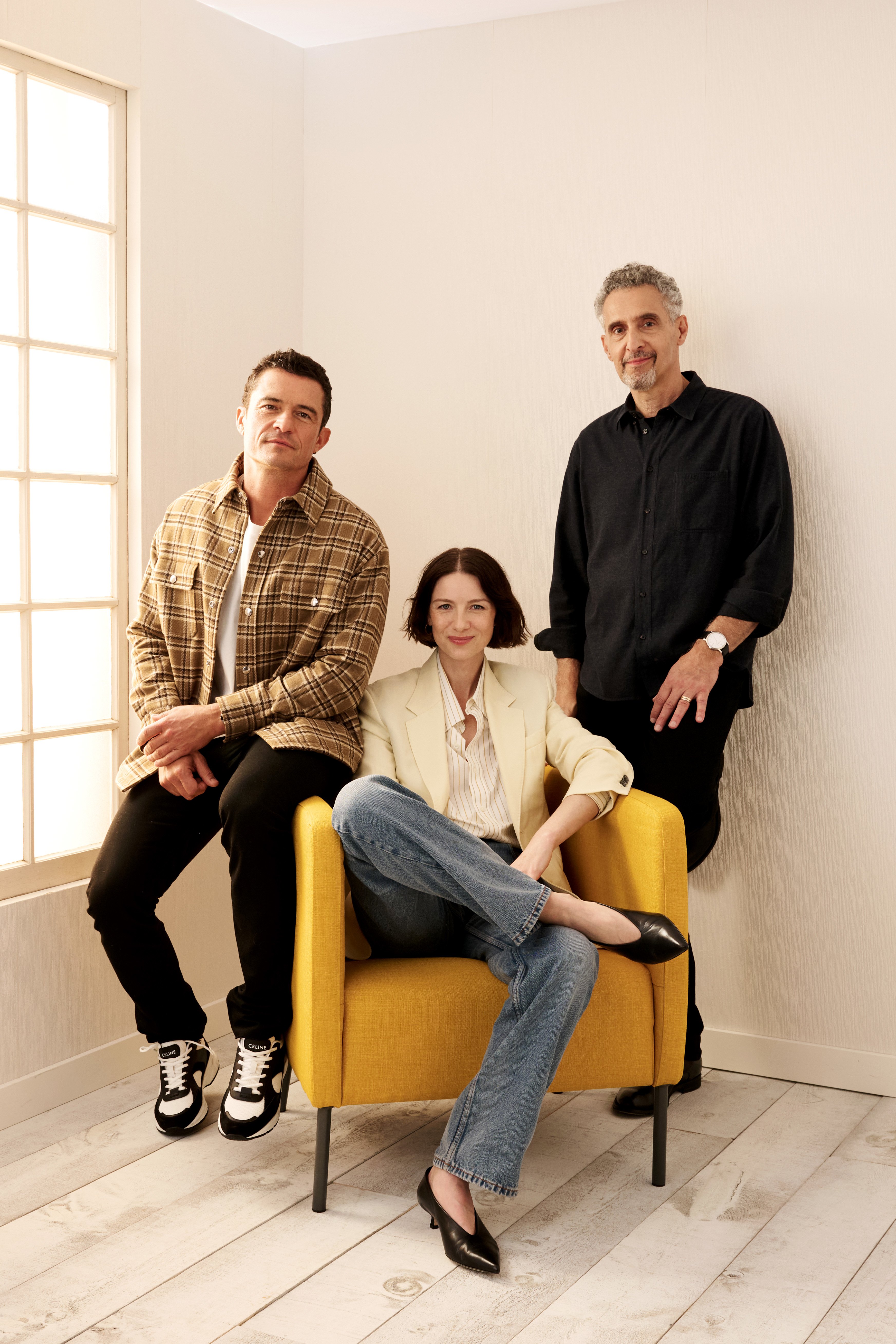 Orlando Bloom, Caitriona Balfe, and John Turturro pose in a room. Hathaway is seated on a yellow chair, wearing jeans and a blazer. Brody and Turturro stand beside her