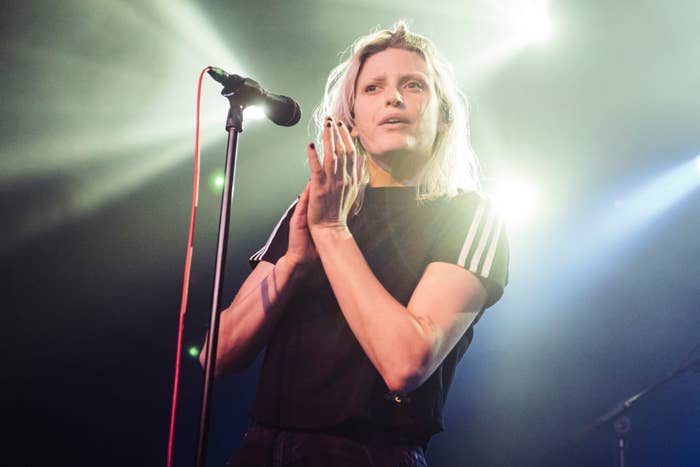 A woman with shoulder-length hair claps her hands while holding a microphone. She is wearing a casual outfit with a striped detail on her sleeves
