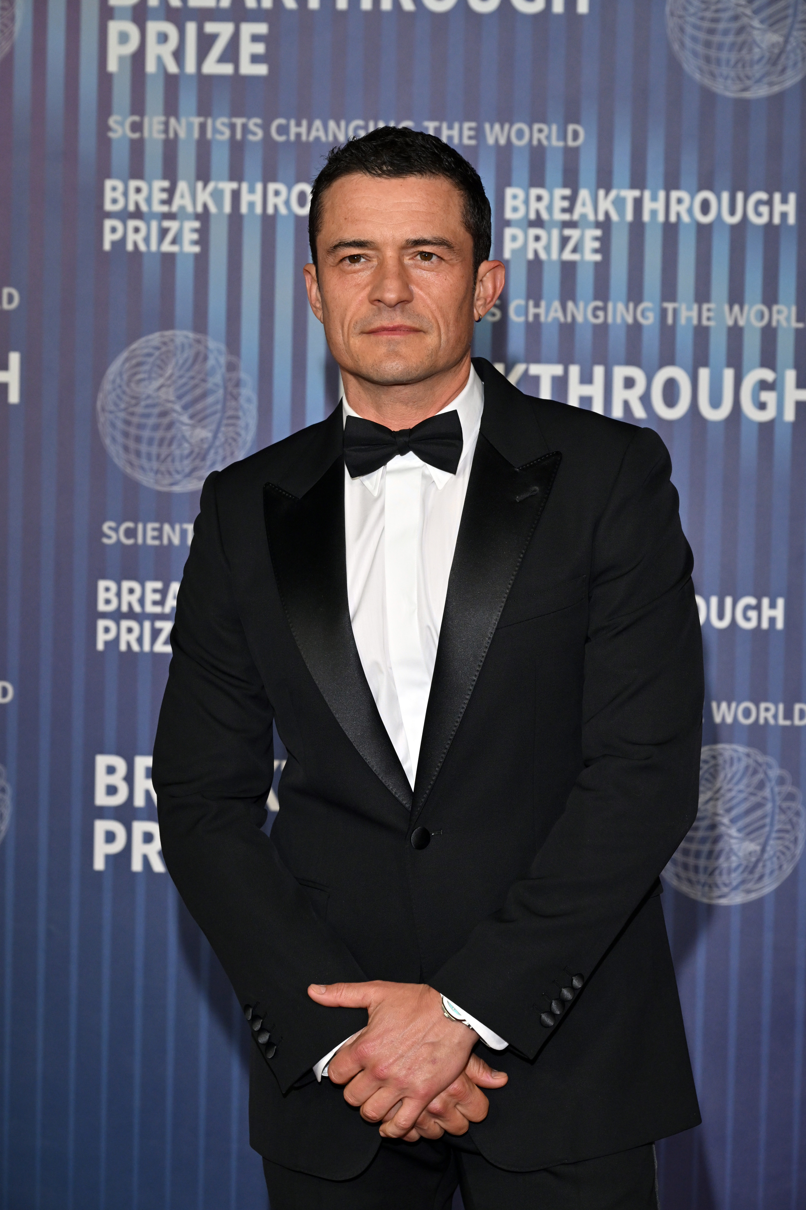 Orlando Bloom at the Breakthrough Prize event, wearing a black tuxedo with a bow tie, standing in front of a backdrop with event branding
