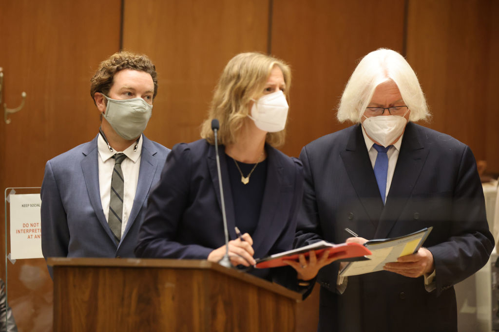 Danny Masterson in a suit and mask stands beside two attorneys in a courtroom, all wearing masks. A sign in the background with legal text is partially visible