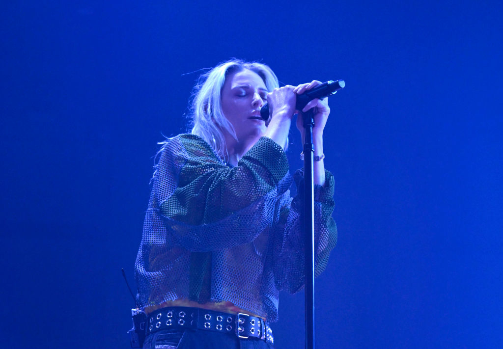 Billie Eilish singing on stage, wearing a loose mesh top and belted pants