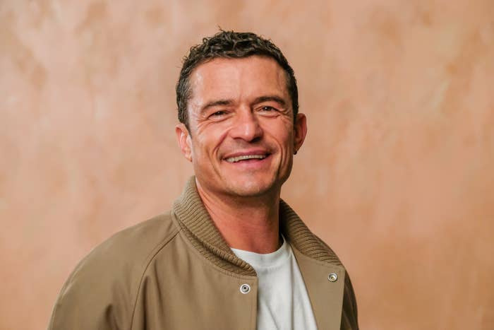 Orlando Bloom smiles, wearing a white t-shirt and a casual beige jacket