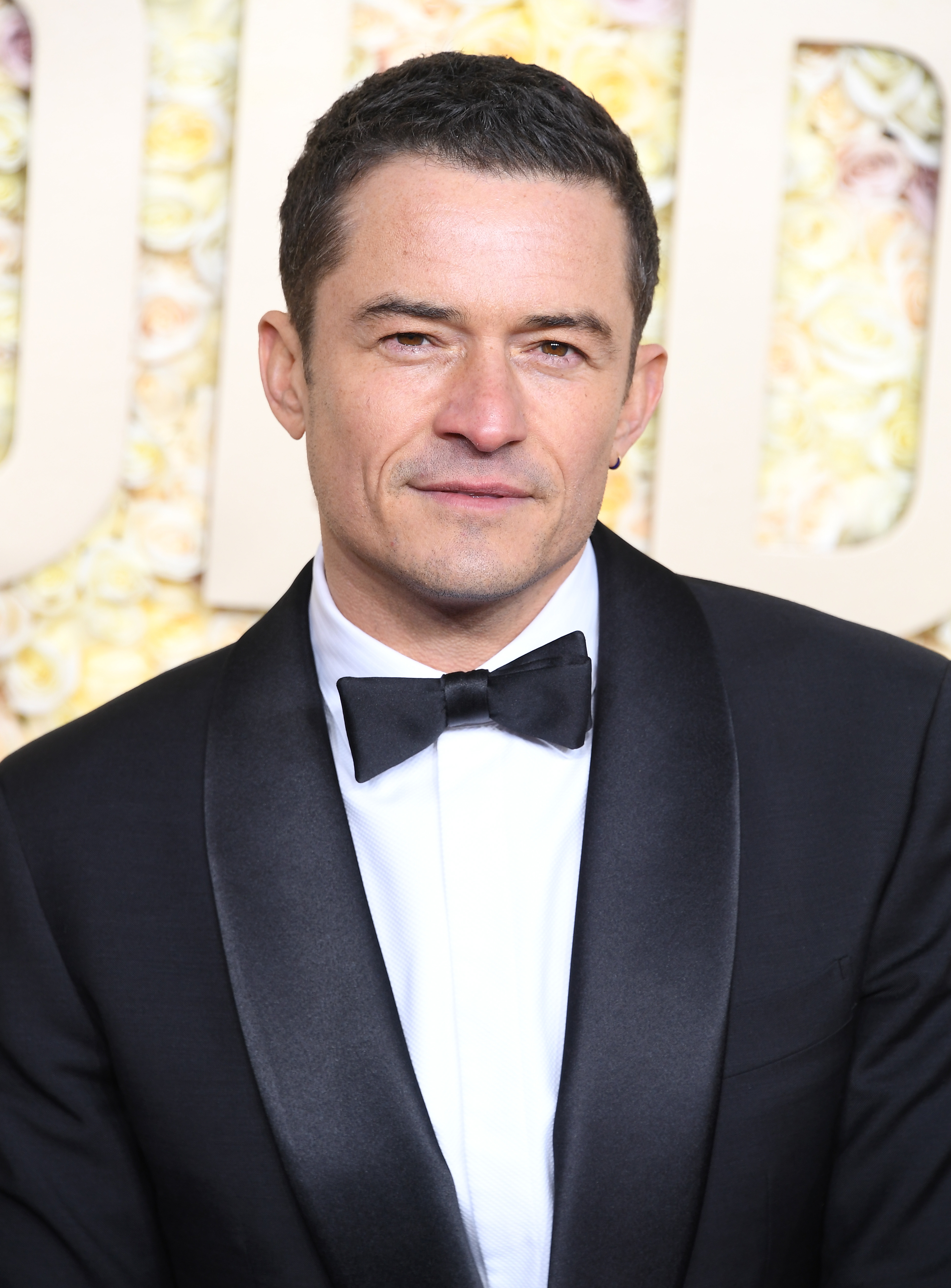Orlando Bloom is dressed in a sharp, black tuxedo with a black bow tie, standing against a backdrop with floral elements and the letters &quot;OD&quot; in the background