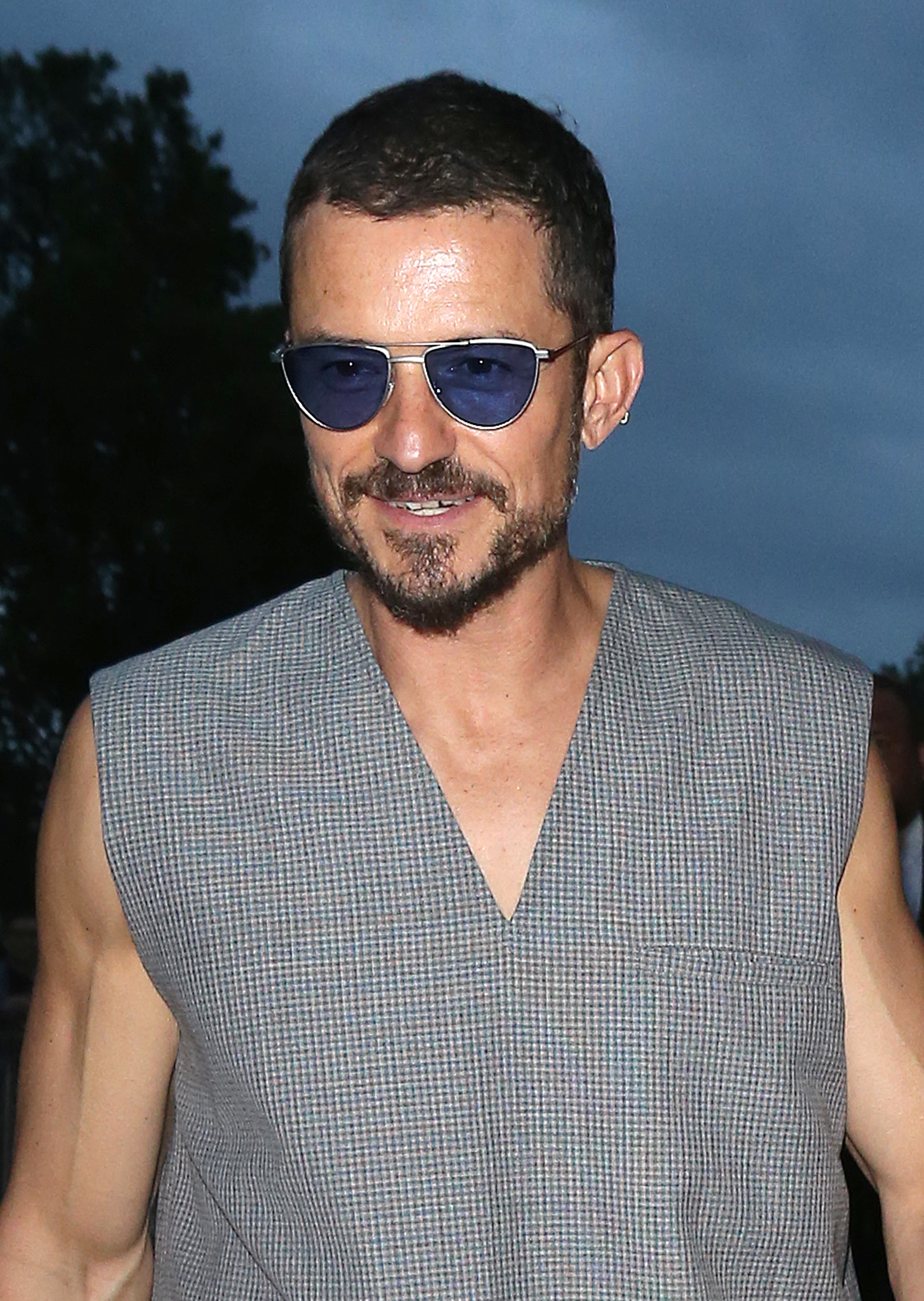 Orlando Bloom smiles, wearing sleeveless, V-neck gray checked shirt and blue-tinted sunglasses