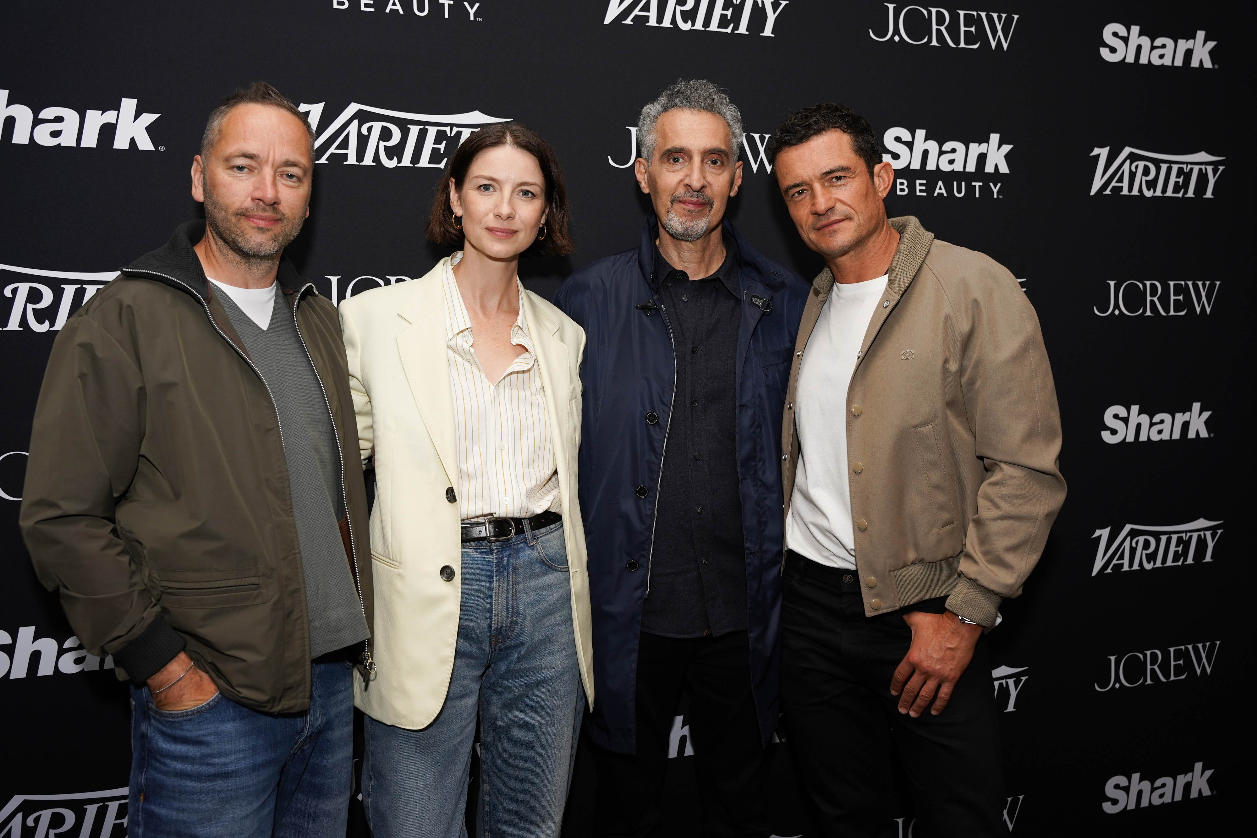 Gavin Hood, Caitriona Balfe, John Turturro, and Orlando Bloom at a Variety event, standing together in casual and semi-formal clothing against a sponsor backdrop