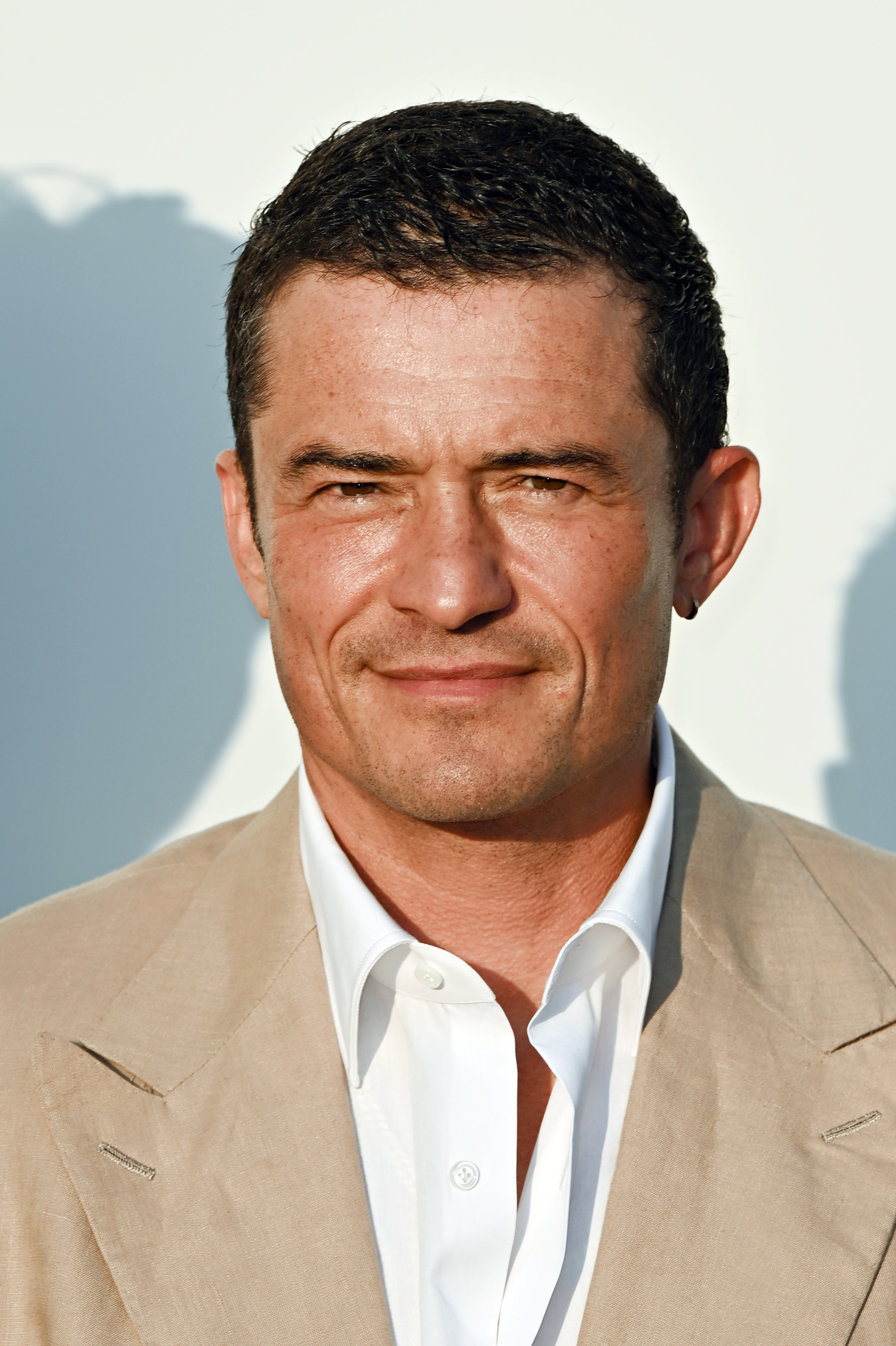 Orlando Bloom smiles, wearing a beige suit jacket over a white dress shirt, minimal background