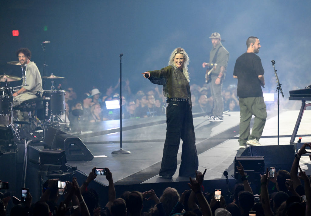 A band performs live on stage with a lead female vocalist, guitarist, drummer, and keyboardist. The audience watches and records the show with their phones