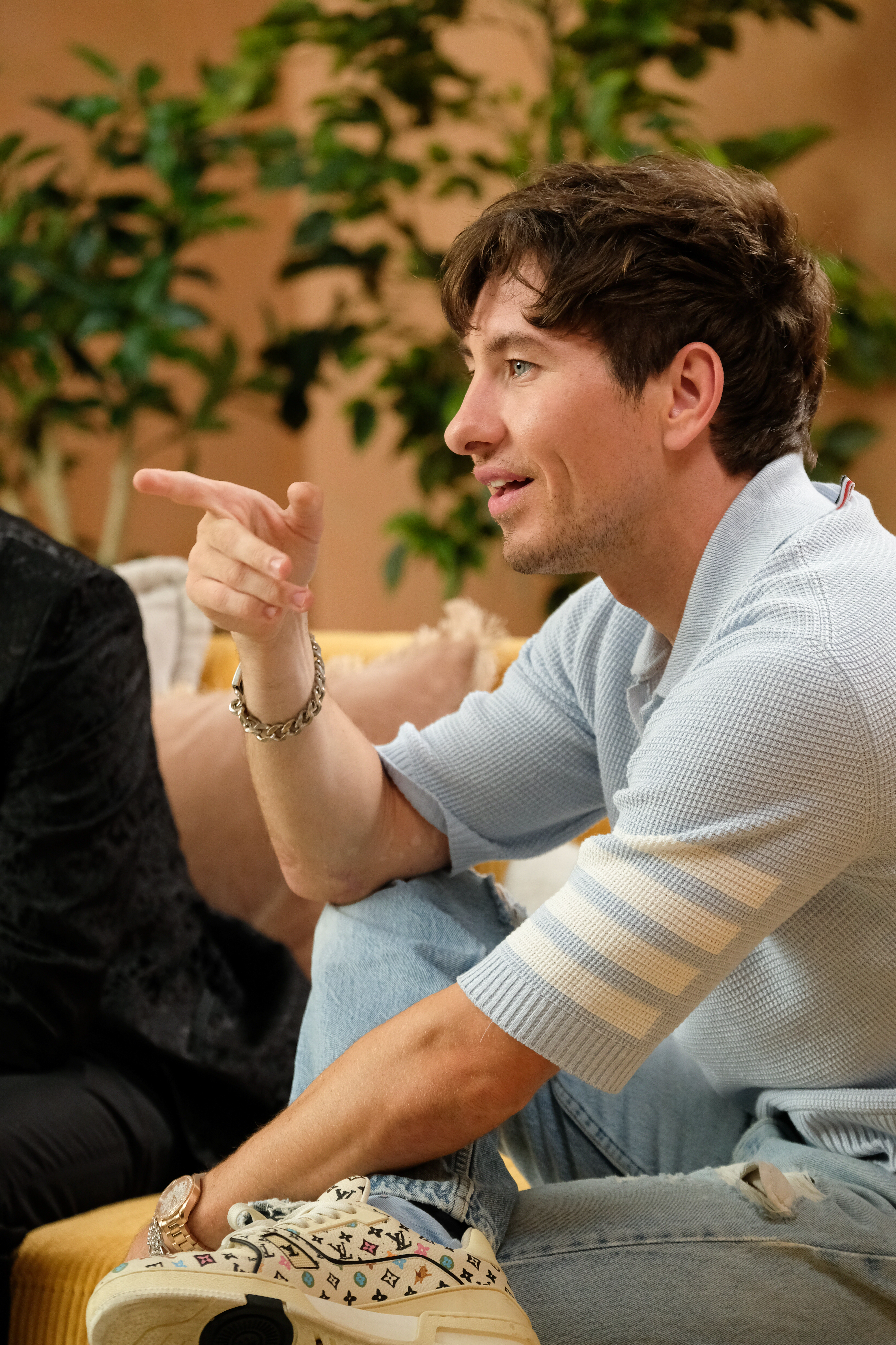 Barry Keoghan is sitting on a couch, wearing a light shirt and jeans, pointing while engaging in a conversation