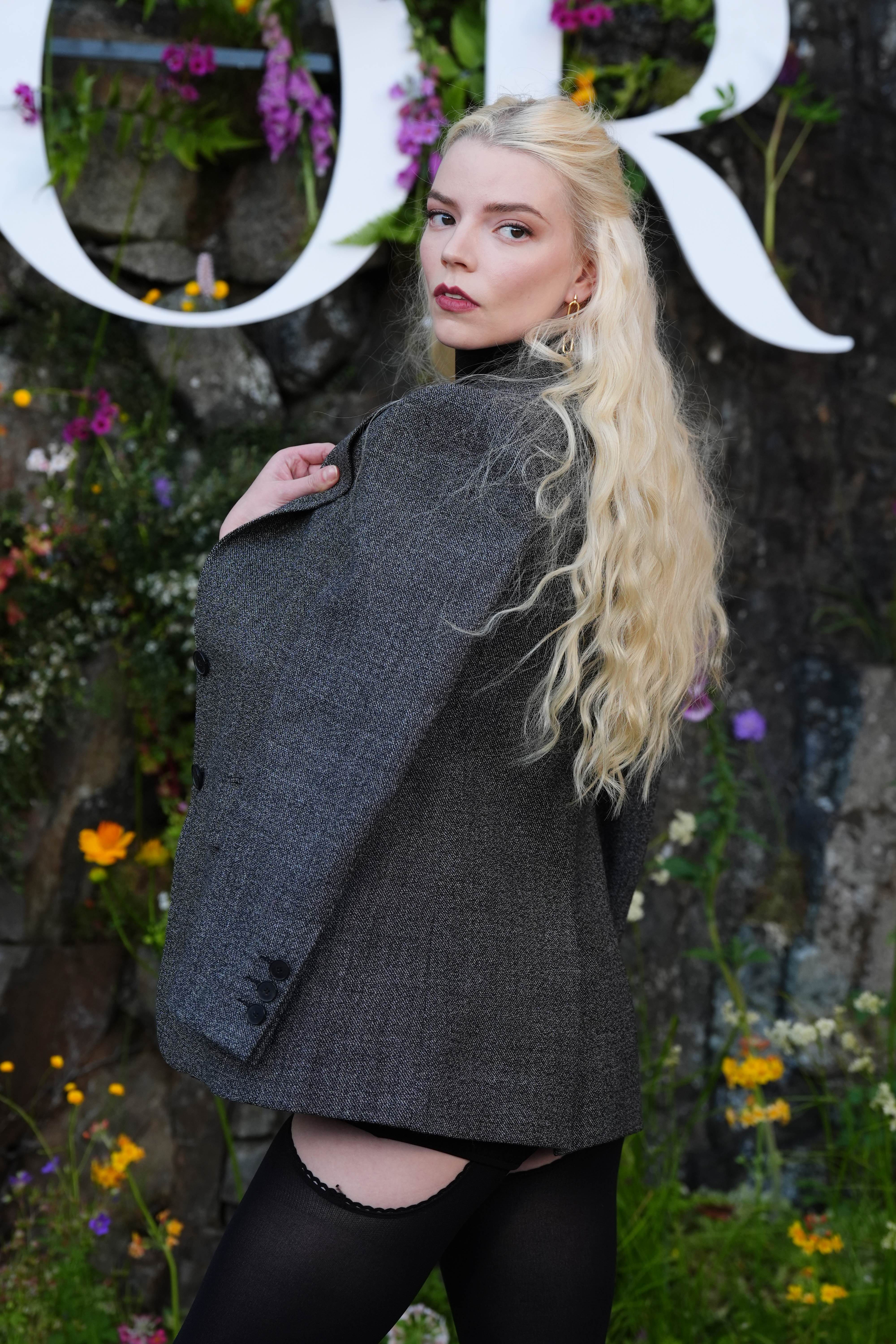 Anya Taylor-Joy poses outdoors in a grey blazer and stockings, with long wavy hair cascading down her back. Floral decorations are in the background