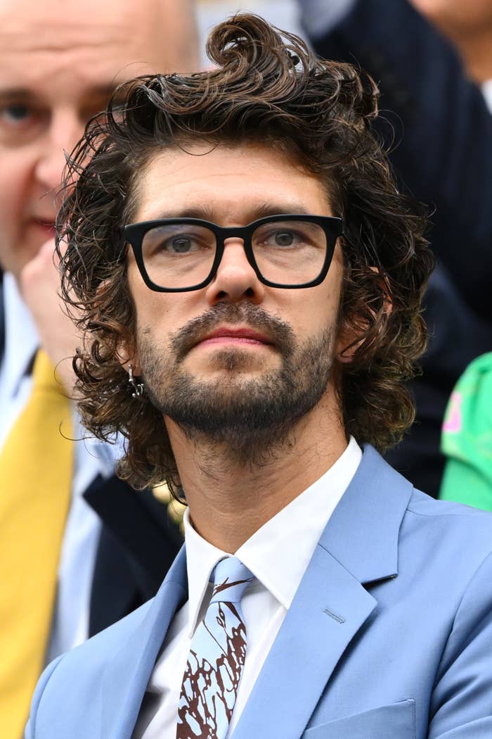 Ben Whishaw wearing a light suit, patterned tie, and glasses, looking slightly upward. A person in the background is partially visible