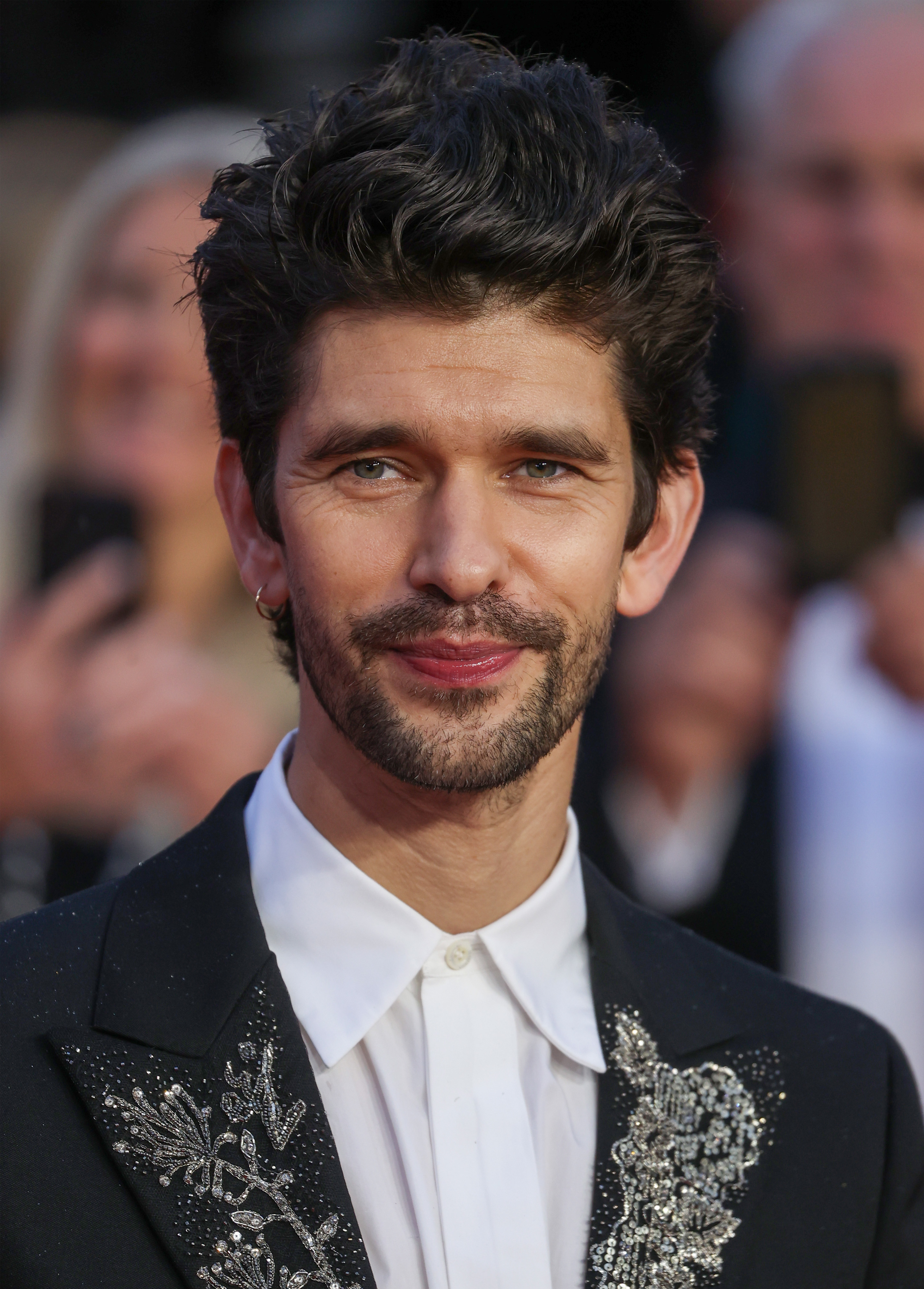 Ben Whishaw on a red carpet, wearing a suit jacket with ornate floral embroidery, white shirt, and sporting a neatly trimmed beard
