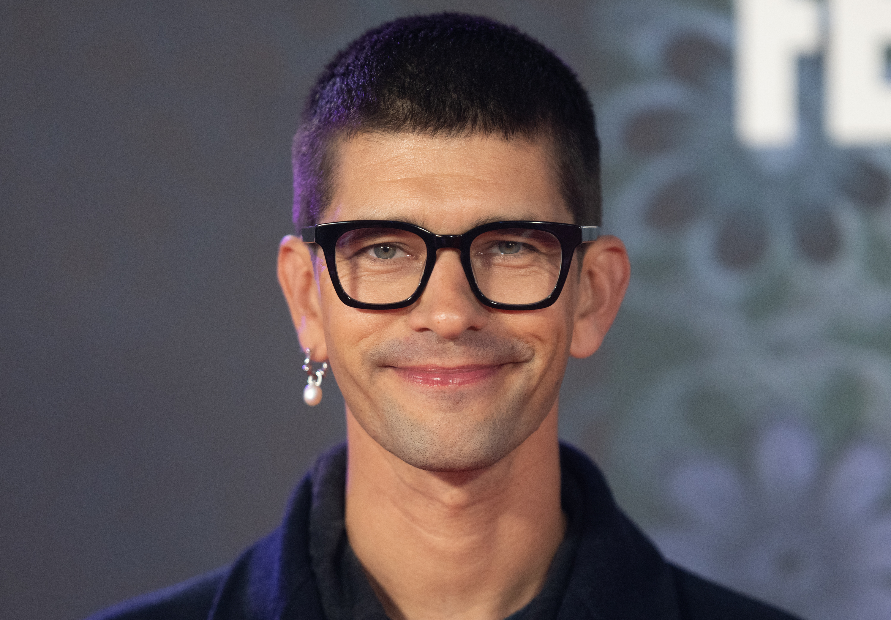 Ben Whishaw smiles at the camera in black-rimmed eyeglasses, a pearl earring, and a dark outfit