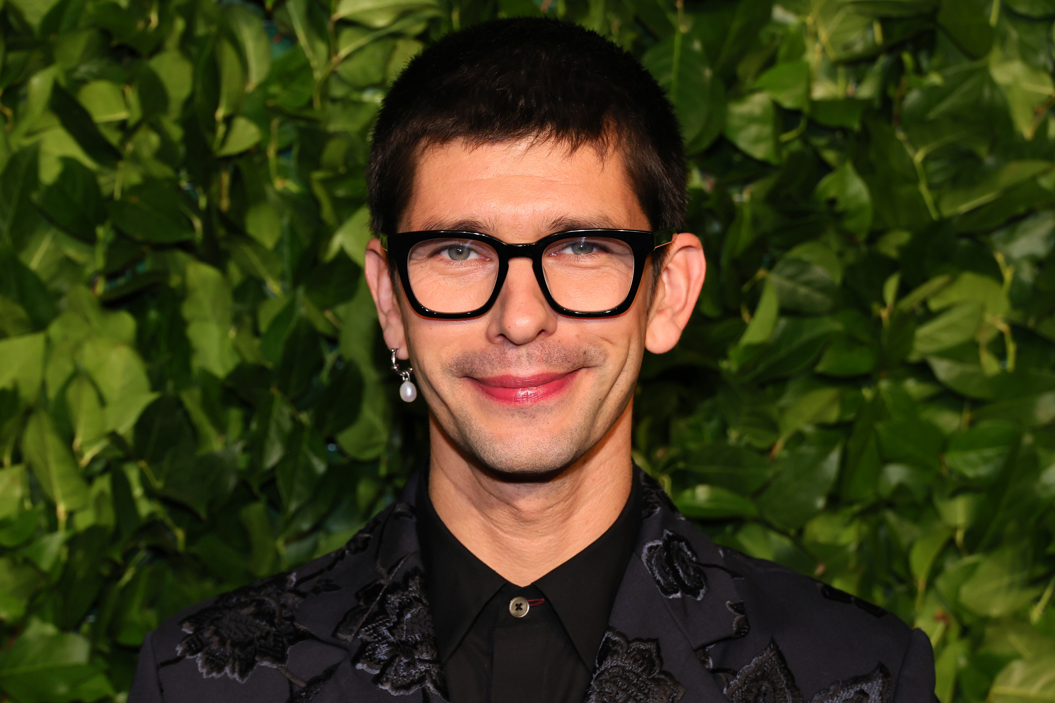 Person smiling in front of a leafy backdrop, wearing glasses and a dark patterned jacket