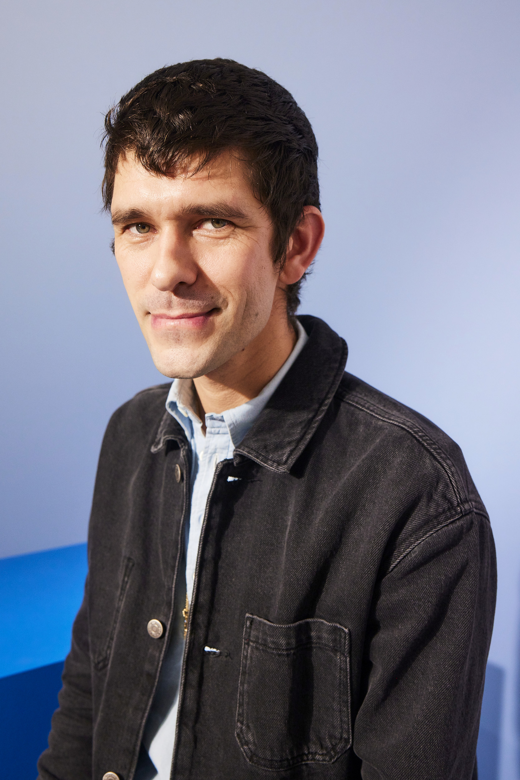 Ben Whishaw is shown in a casual portrait, wearing a light shirt and a dark denim jacket, smiling at the camera. Background is a plain setting
