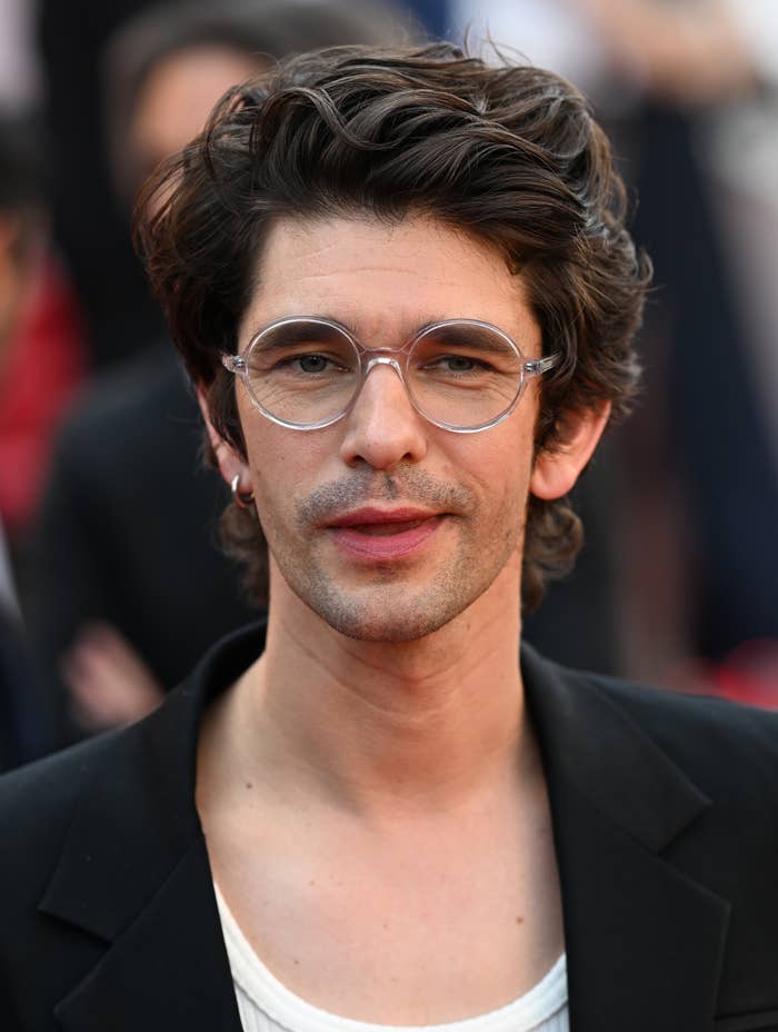 Person with medium-length wavy hair and glasses, wearing a white shirt under a black jacket on a red carpet