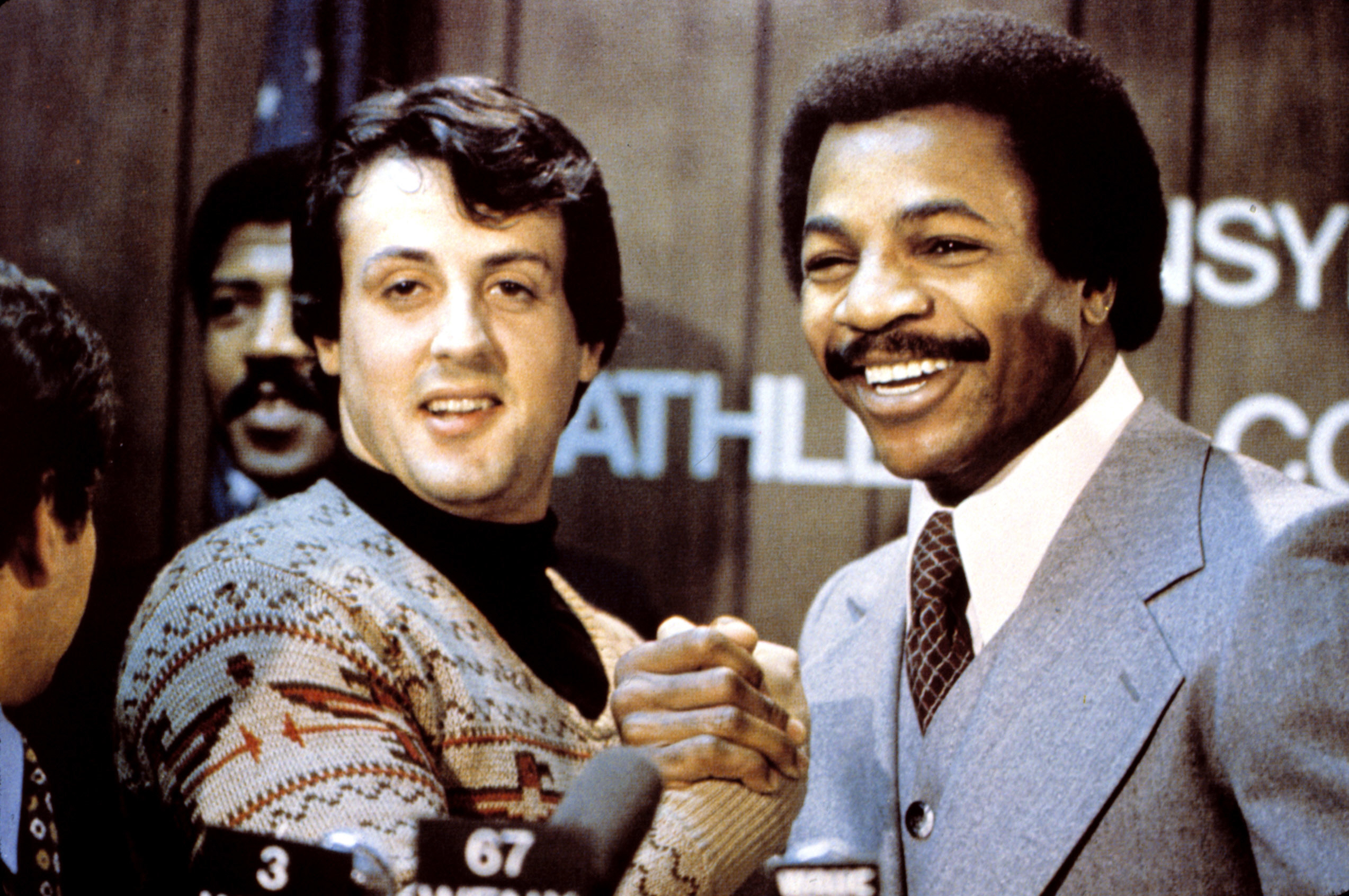 Sylvester Stallon shaking hands with Carl Weathers in a photo opp in "Rocky"