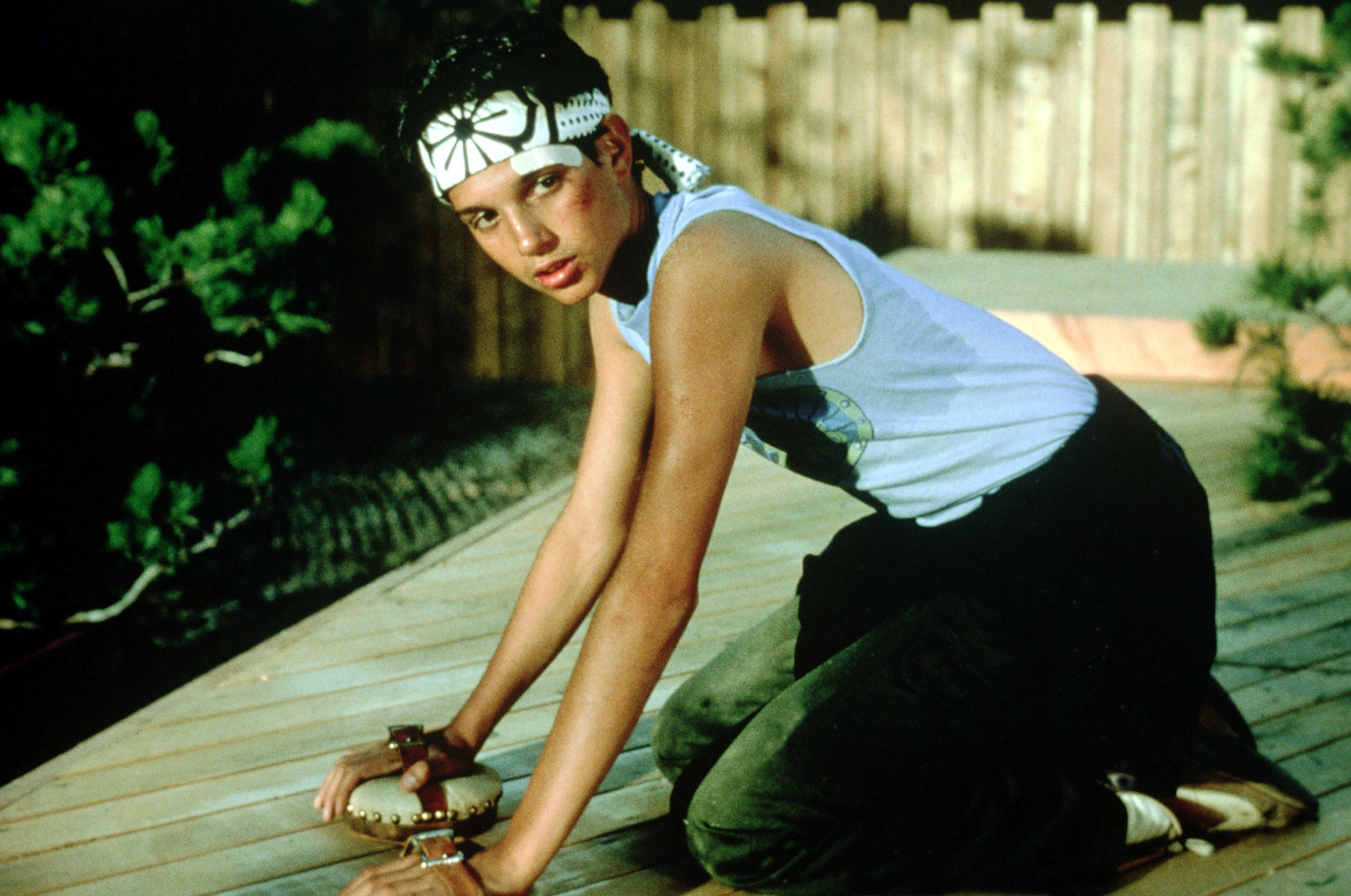 Ralph Macchio, in a headband and tank top, kneels while waxing a wooden deck in a scene from "The Karate Kid"