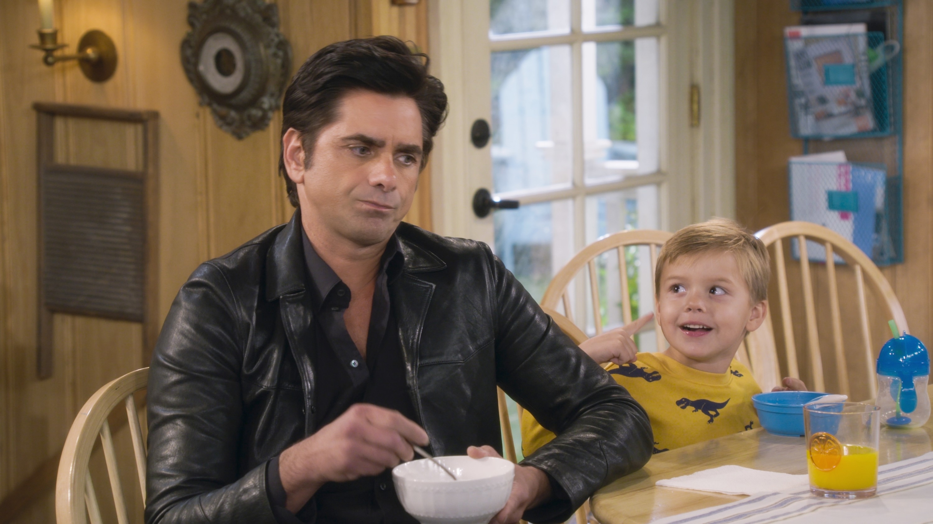 John Stamos sits at a kitchen table eating cereal next to a kid in "Fuller House"