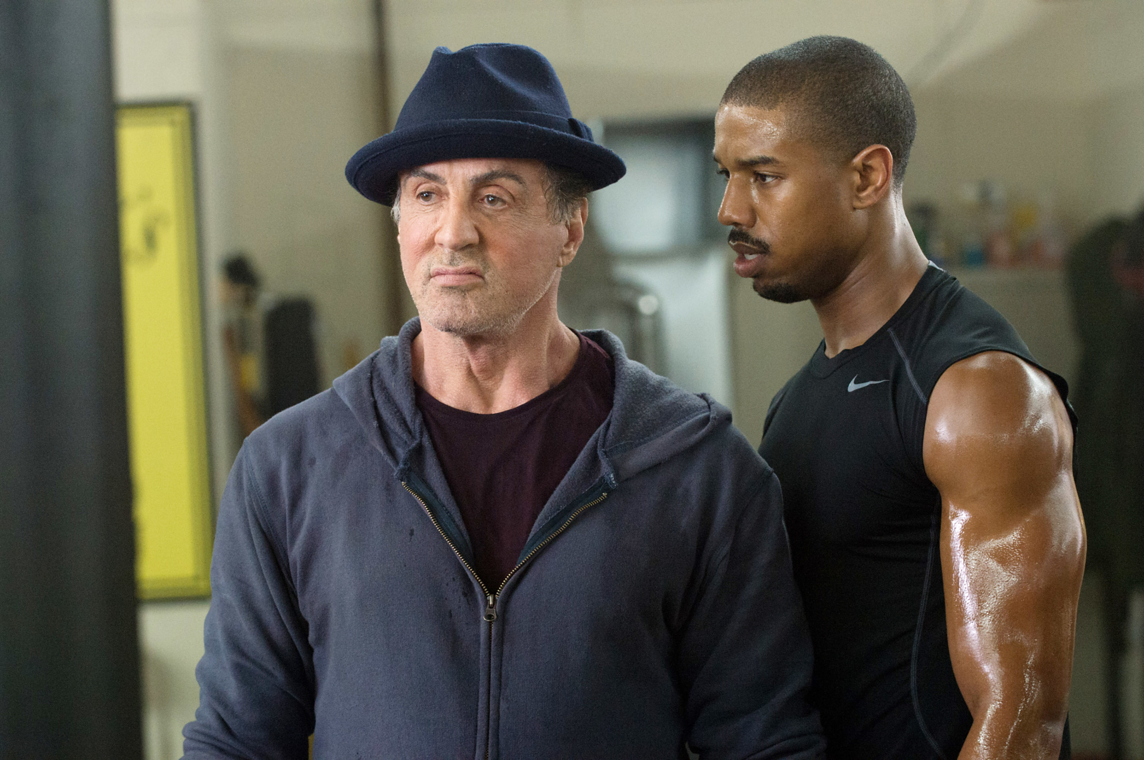 Sylvester Stallon wearing casual clothing and a fedora as he stands next to Michael B. Jordan in "Creed"