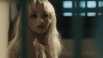 Woman with blonde hair and bold makeup looking to the side through a set of bars, in a scene from a music video