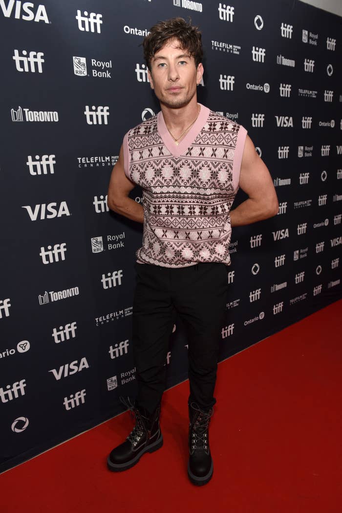 Ezra Miller poses on the red carpet at TIFF, wearing a patterned sleeveless sweater, black pants, and lace-up boots