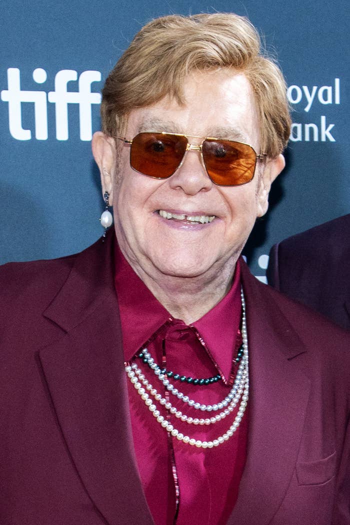 Elton John on the red carpet at the Toronto International Film Festival, wearing a burgundy suit with layered pearl necklaces and orange-tinted glasses