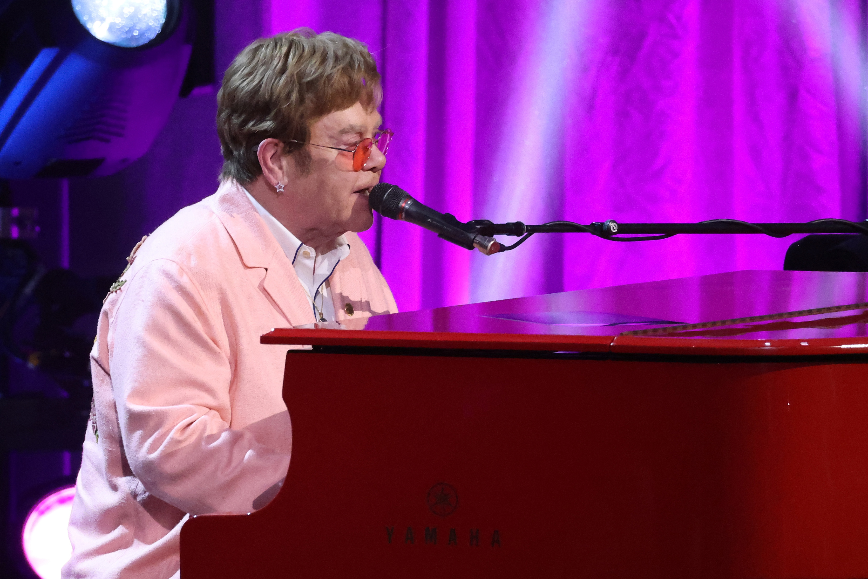 Elton John plays a red Yamaha piano while singing into a microphone, dressed in a pink blazer and orange-tinted glasses