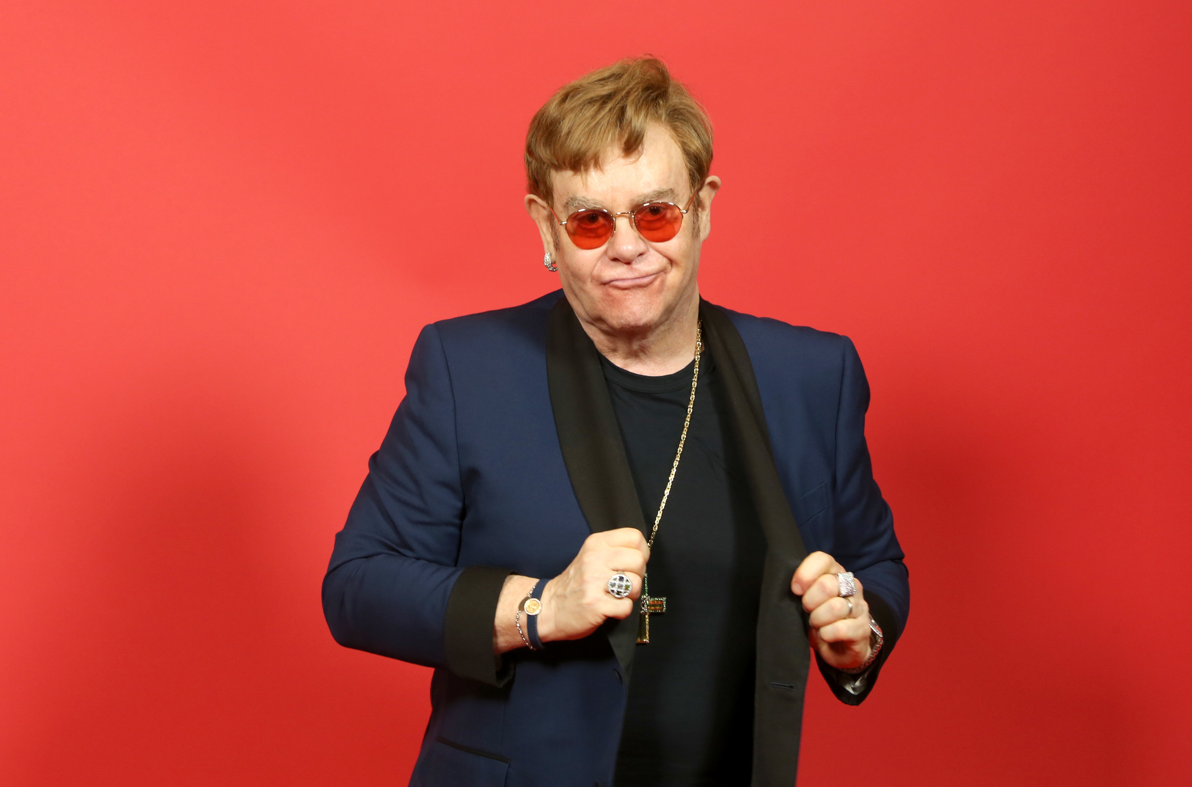 Elton John is posing against a red background, wearing a blue blazer with black lapels, a necklace with a cross, and red-tinted sunglasses