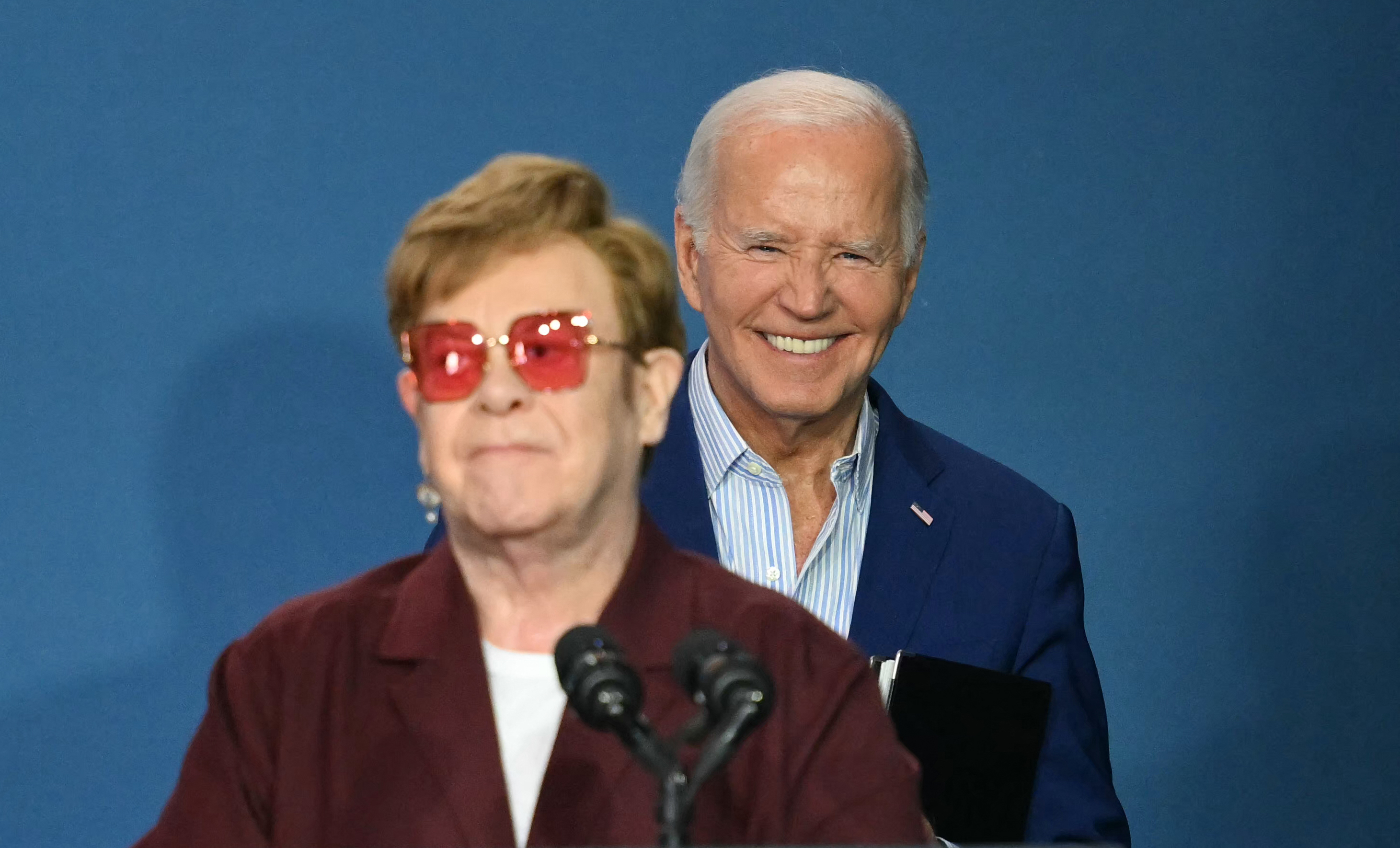 Elton John, in a casual outfit and red-tinted glasses, stands at a microphone, with President Joe Biden smiling in the background