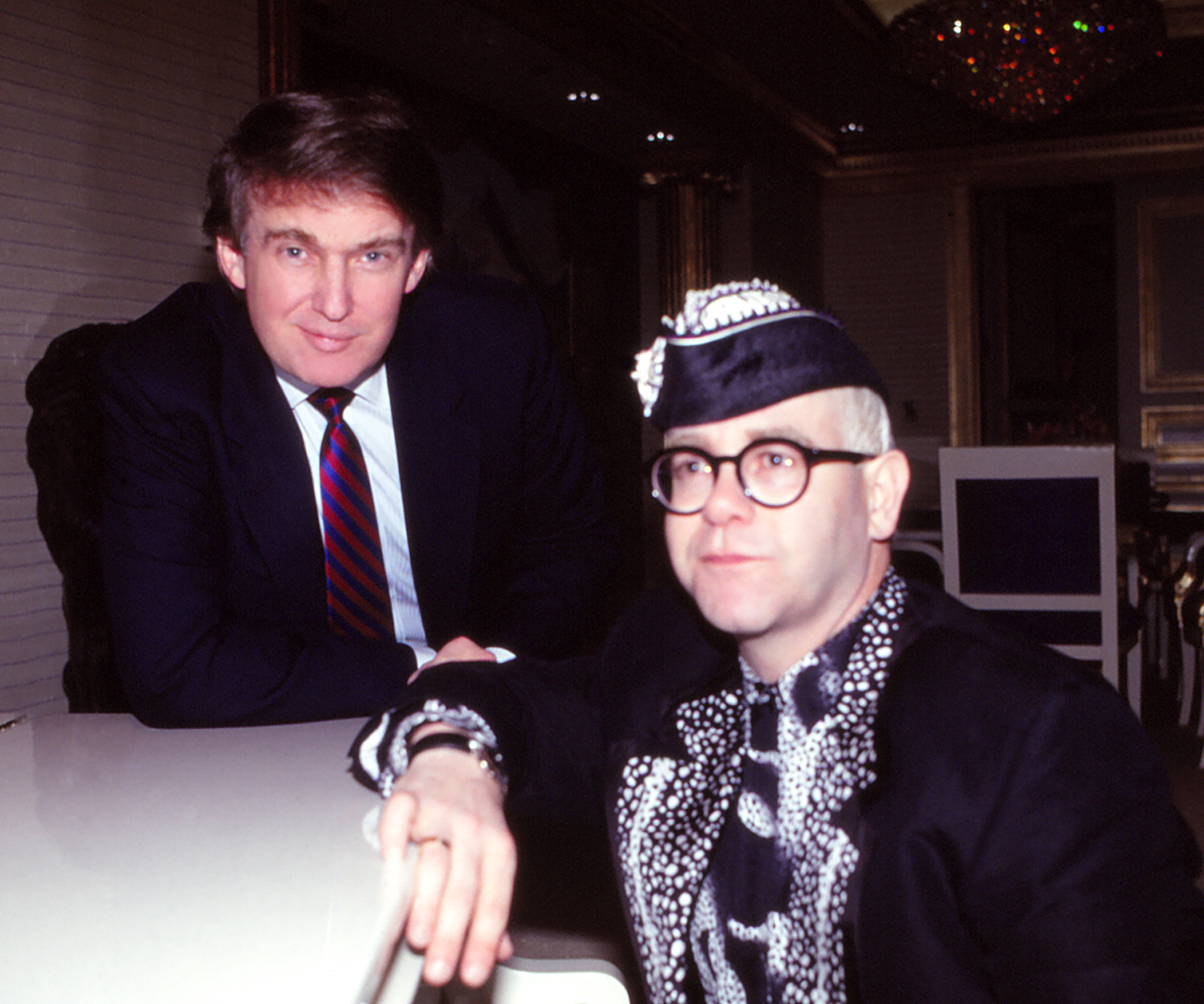 Donald Trump and Elton John pose together, with Trump wearing a suit and tie, and John dressed in a patterned outfit with glasses and a hat