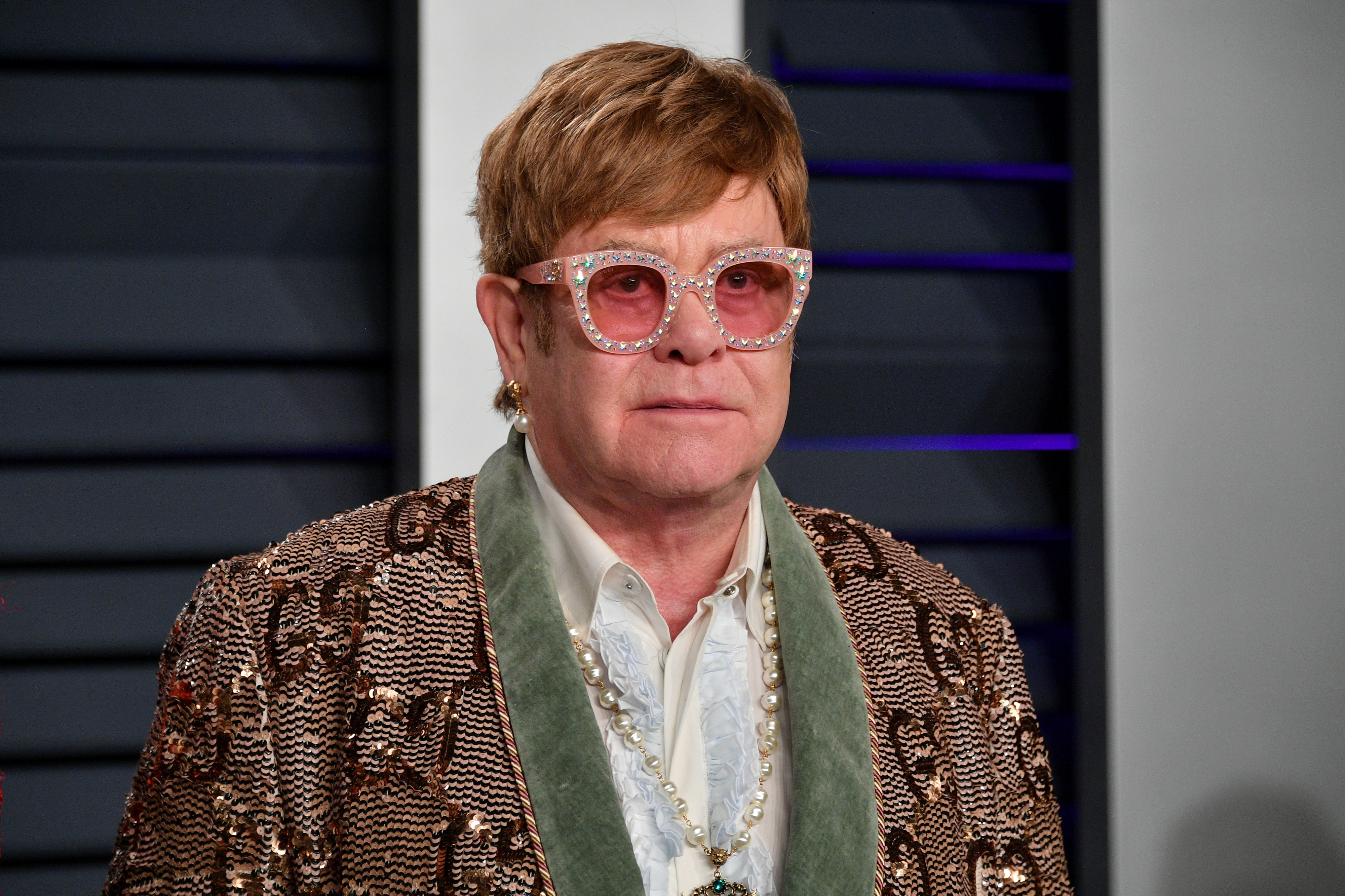 Elton John wears ornate glasses and a detailed, patterned blazer with a pearl necklace at a red carpet event