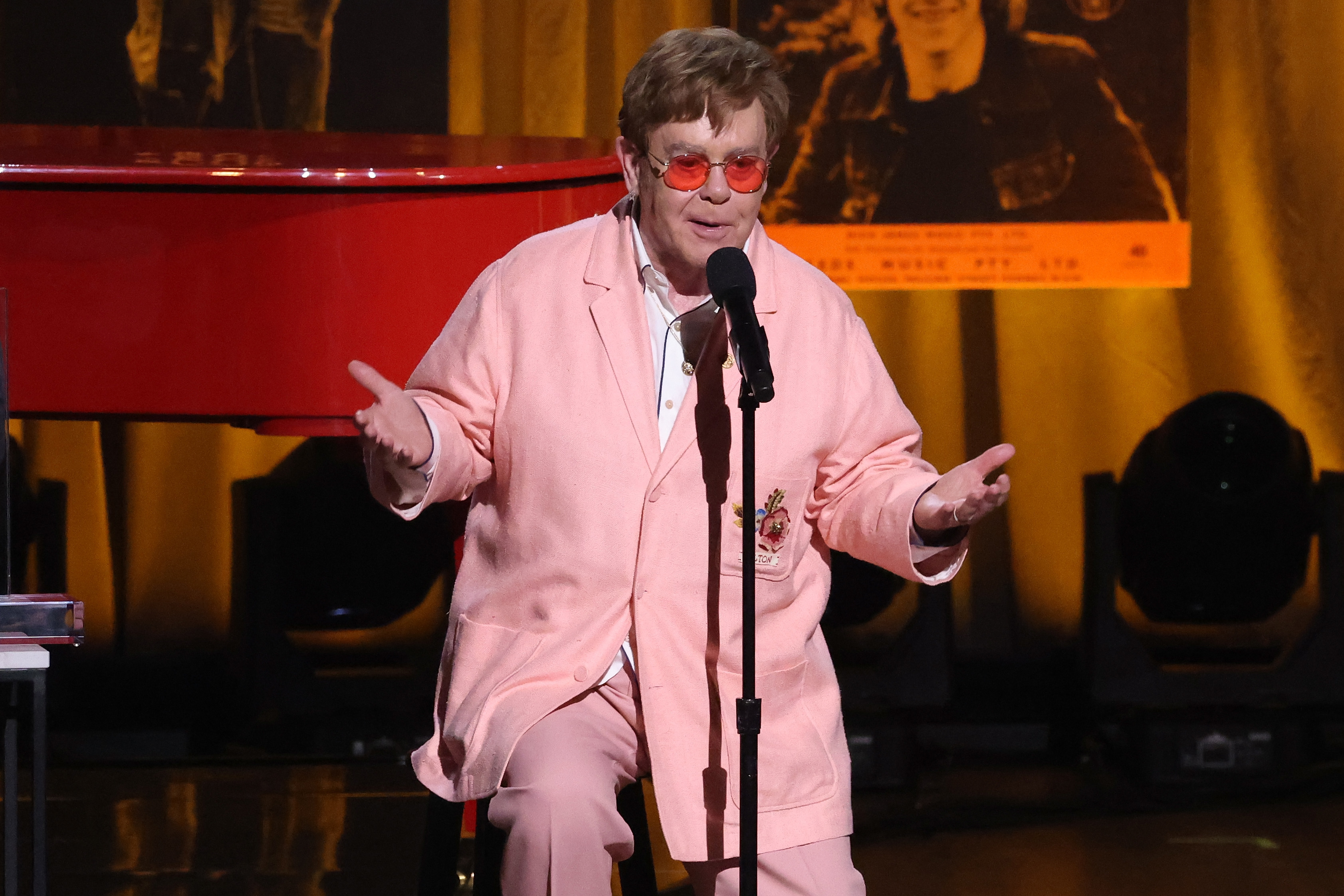 Elton John speaks into a microphone while wearing a pink jacket and pants with red-tinted glasses, performing on stage next to a red piano