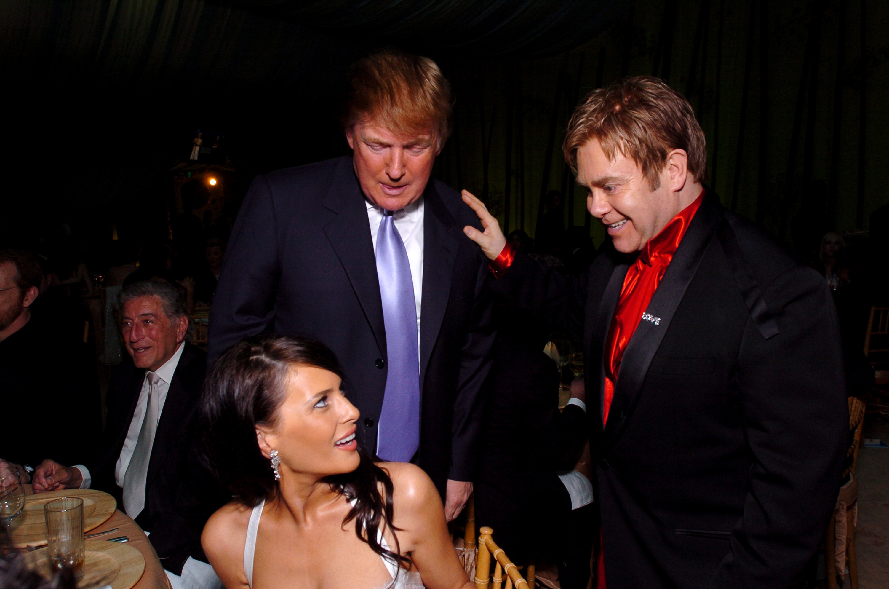 Donald Trump stands and speaks with seated woman and Elton John, who is wearing a suit with a red scarf, at an event