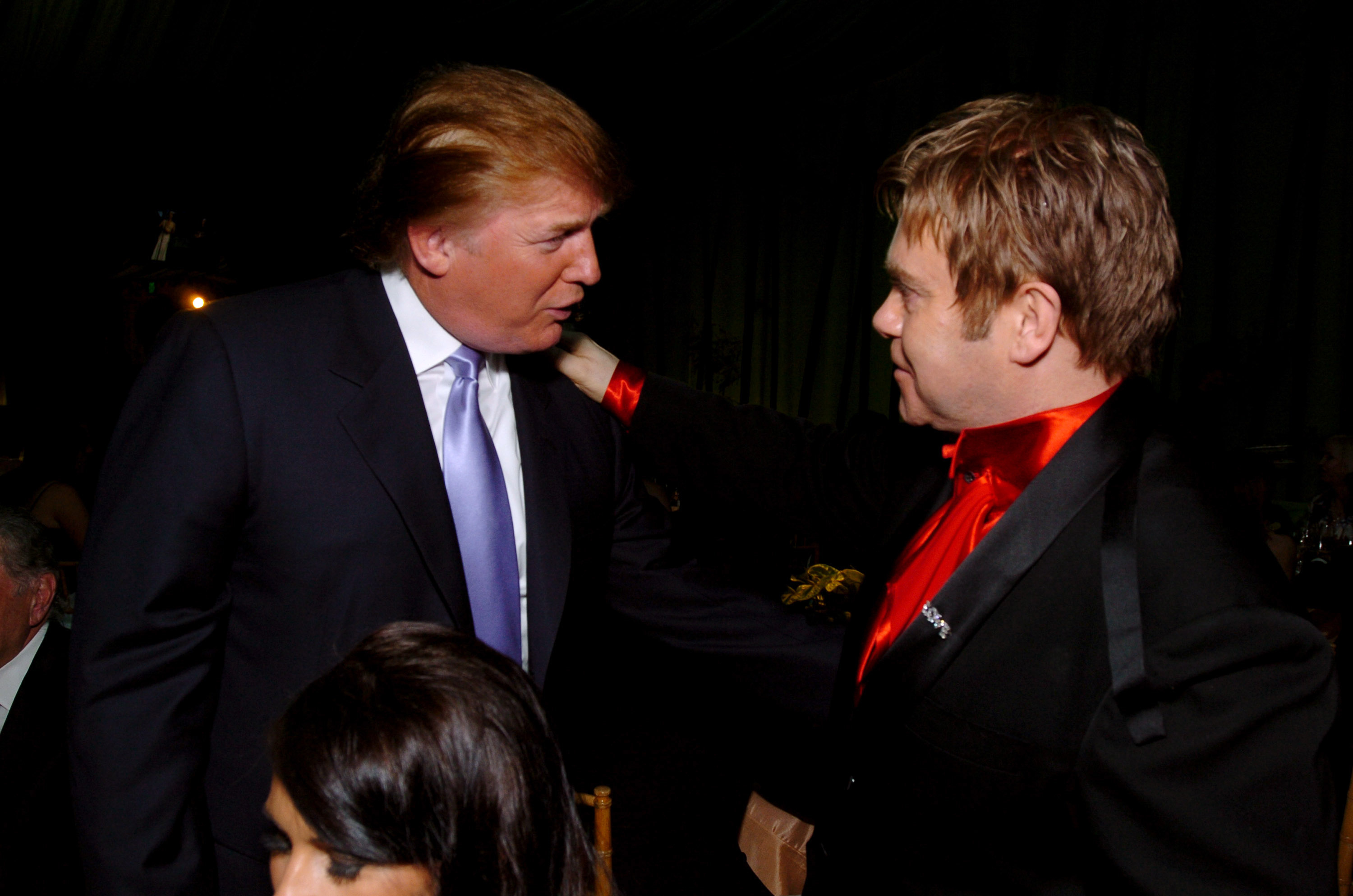 Donald Trump in a suit and tie converses with Elton John, who is dressed in a black suit with a red satin shirt